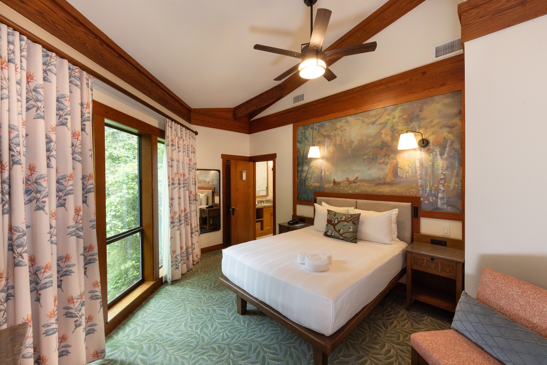 Saratoga Springs new DVC villas.. The image depicts a well-furnished hotel room featuring a double bed with white linens, an artwork on the wall, and large windows overlooking a forested area. The room is characterized by rustic wooden accents and light-colored floral curtains.