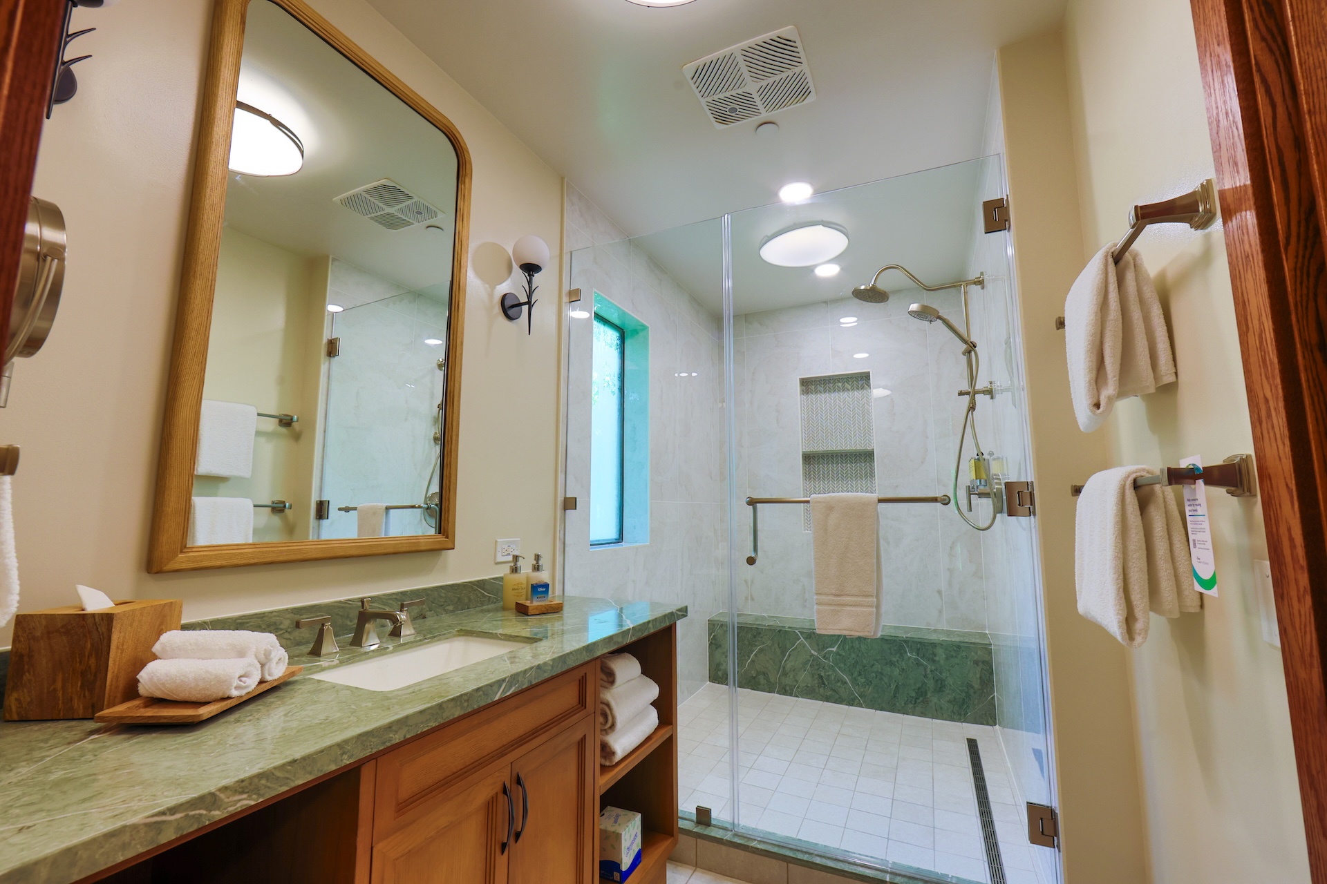Saratoga Springs new DVC villas. The image shows a modern bathroom featuring a large walk-in shower with a glass partition. The decor includes a green marble countertop, wooden cabinetry, and soft lighting, creating a serene atmosphere.