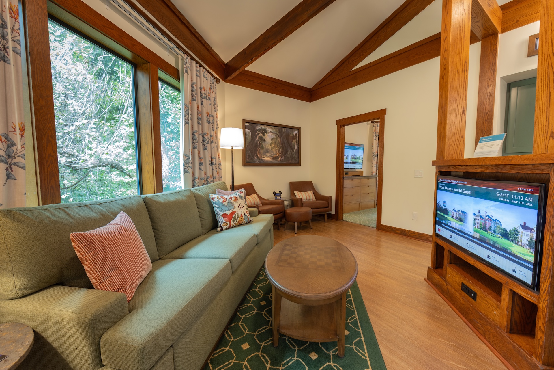 Saratoga Springs new DVC villas. The image depicts a cozy living room featuring a green sofa with decorative pillows, a wooden coffee table, and a large television displaying a property listing. The room is well-lit with natural light filtering through large windows and has wooden beams accentuating the ceiling.