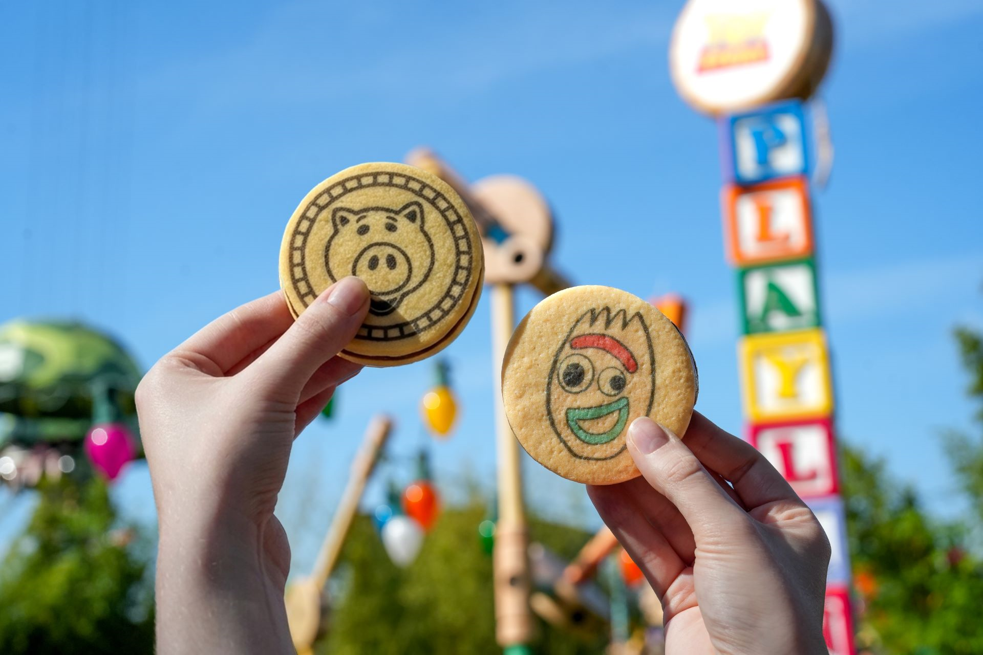 Two hands hold cookies decorated with cartoon characters against a background of colorful blocks and a playful setting. The image captures a fun and cheerful atmosphere, likely intended for children.