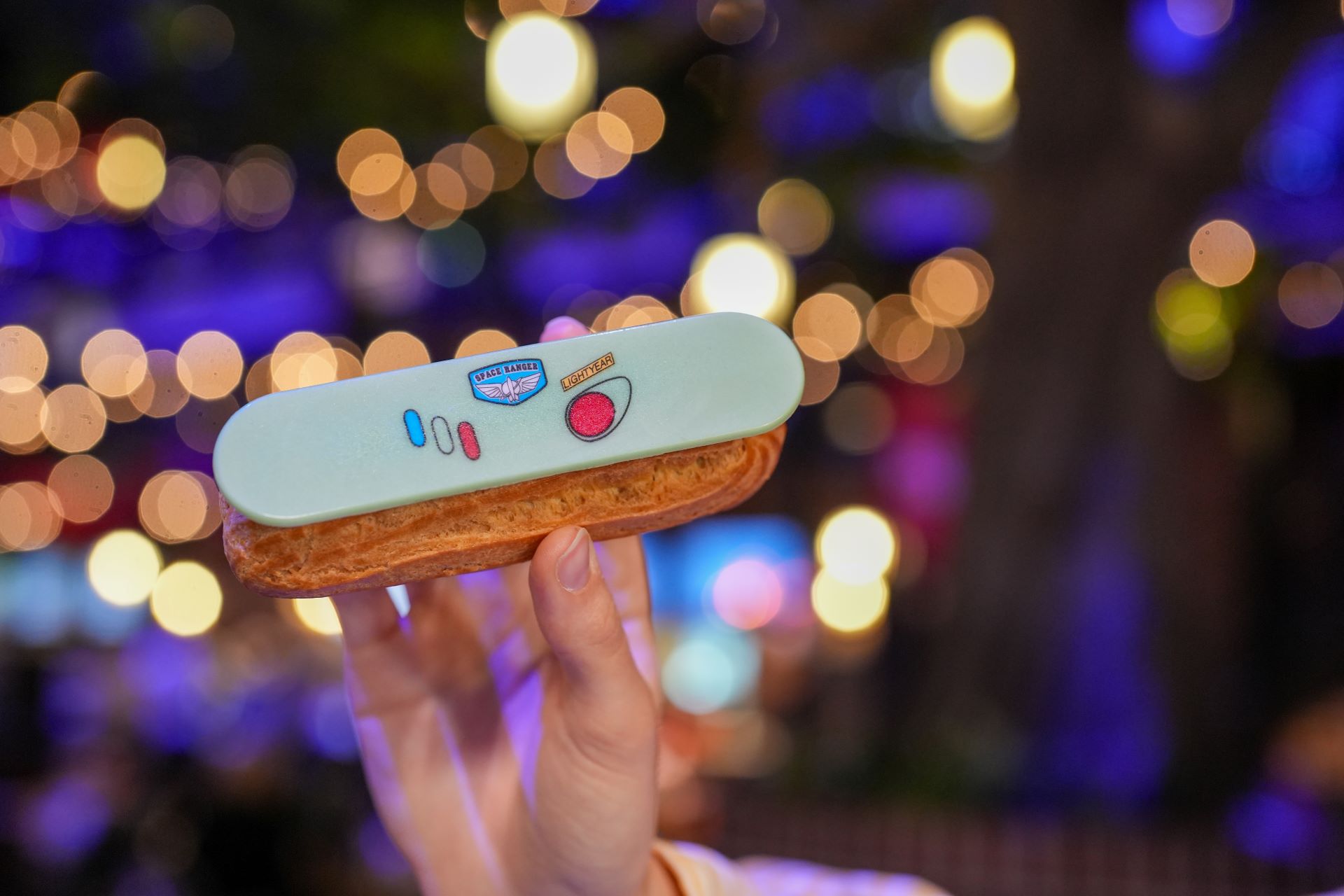 A person holds a decorated pastry featuring colorful designs and a logo, set against a blurred background with warm lights. The image captures a festive ambiance, highlighting the dessert's aesthetic appeal.