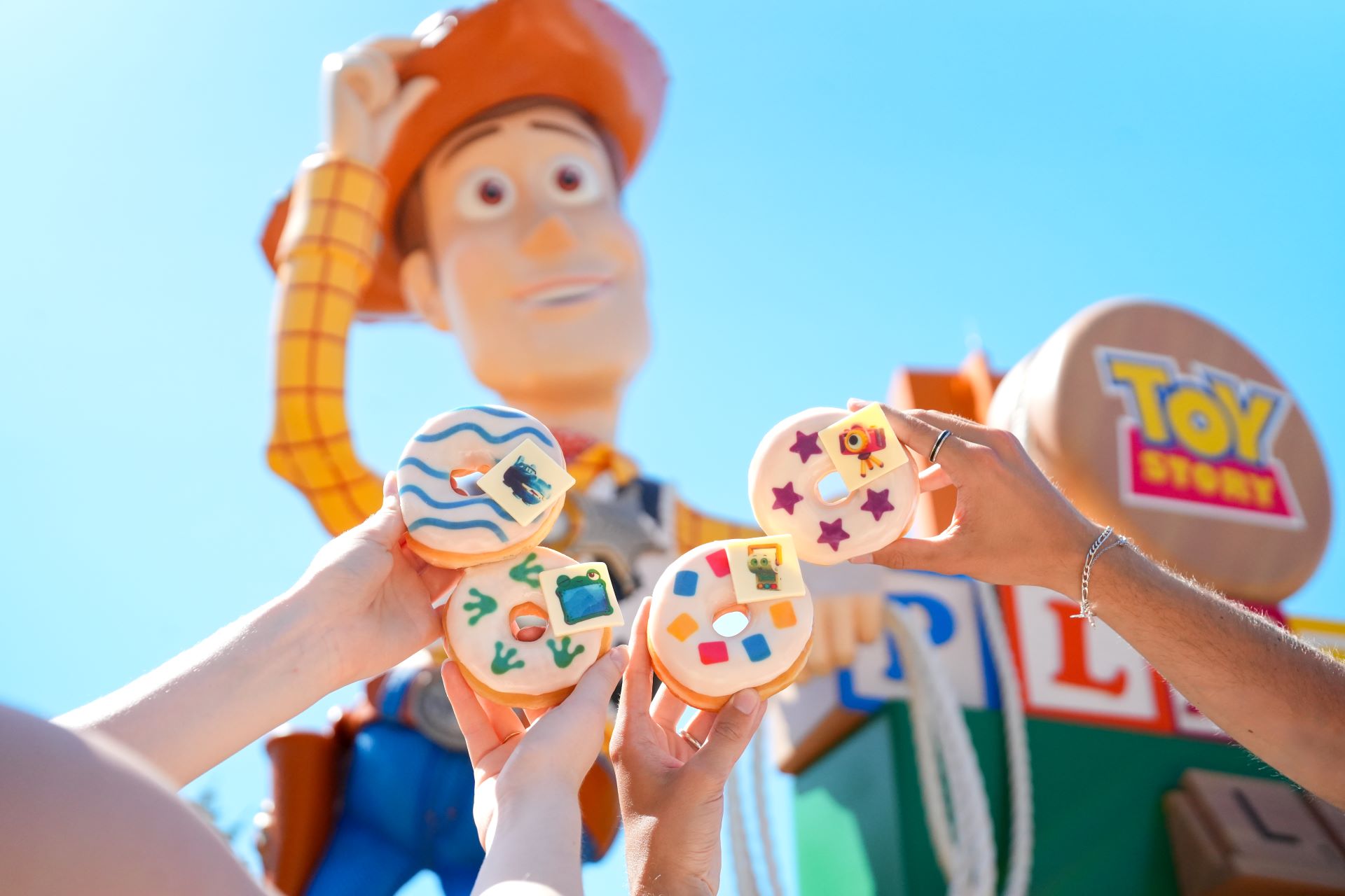 A group of people holds colorful cookies shaped like various characters and items, in front of a large Woody figure from the Toy Story franchise. The scene is set outdoors, under a clear blue sky, highlighting a festive atmosphere.