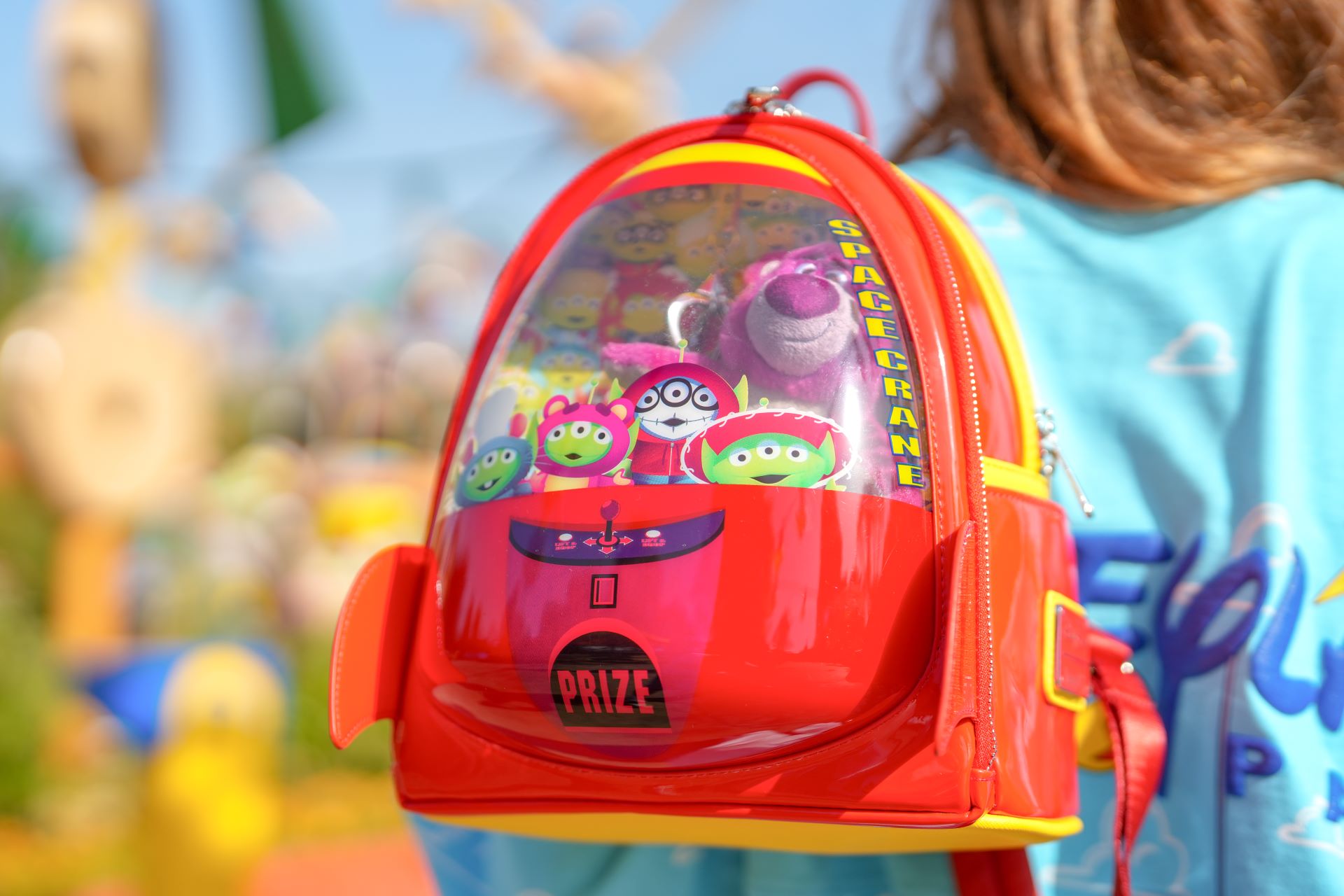 The image features a brightly colored, transparent backpack styled like a toy claw machine. Inside the backpack, cartoon alien figures are visible, suggesting a playful and whimsical design targeted at children.
