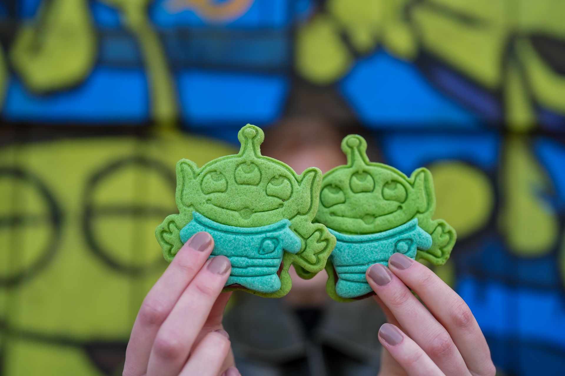 The image features a person holding two green alien-shaped cookies in front of a colorful blue and green graffiti background. The cookies resemble cartoon-like characters, adding a playful and whimsical touch to the scene.