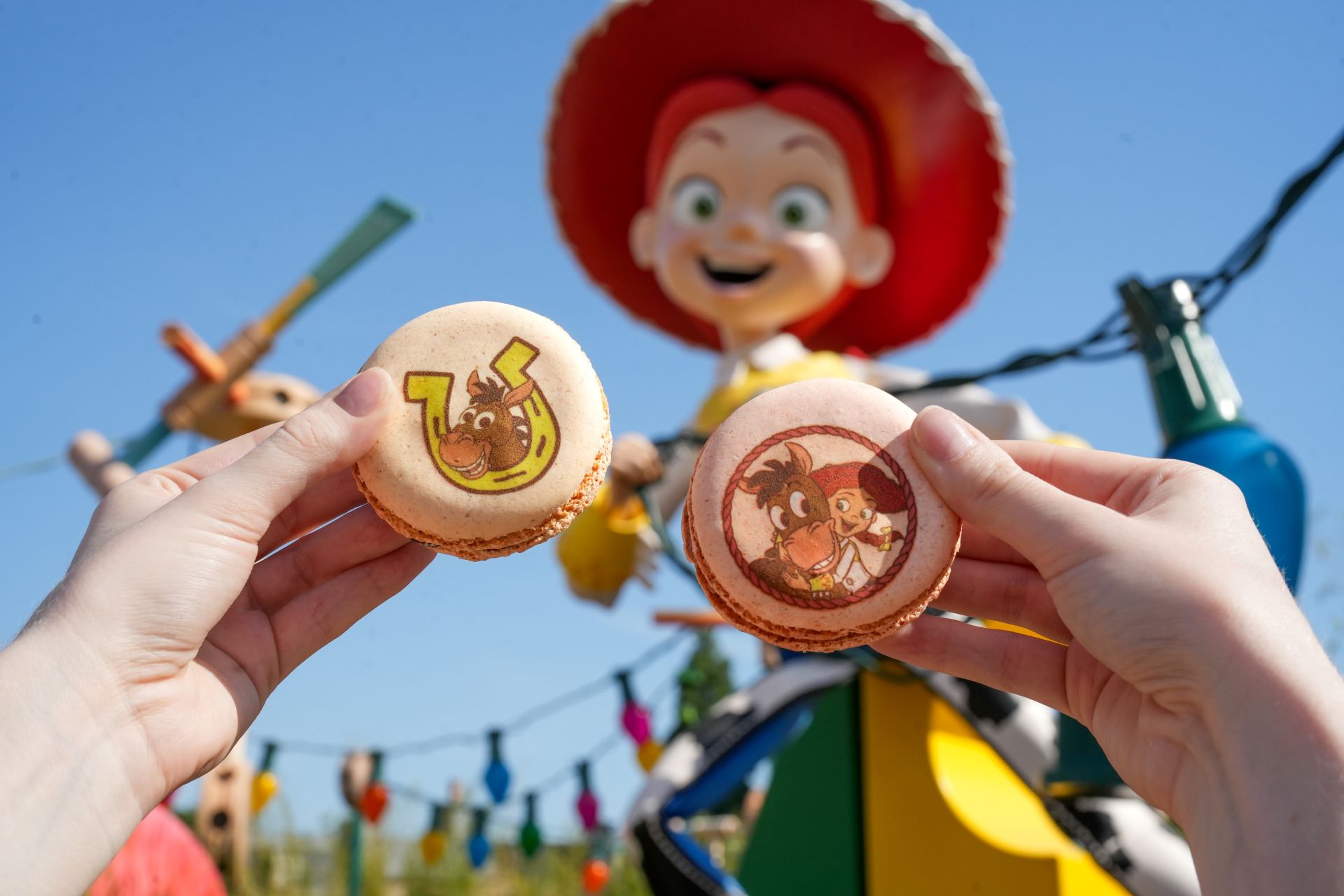 The image features two hands holding macarons with colorful character designs, set against a vibrant outdoor backdrop. In the background, there is a large statue of a character with red hair and a cheerful expression.