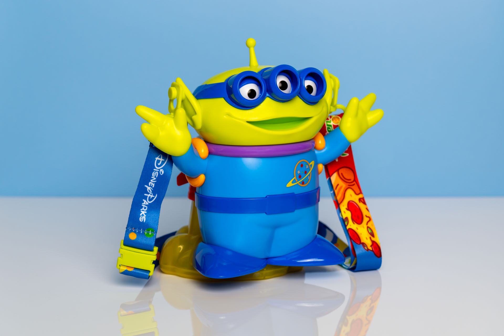 The image features a colorful toy resembling a character with multiple eyes, dressed in a blue outfit. It has vibrant straps with branding from Disney Parks.