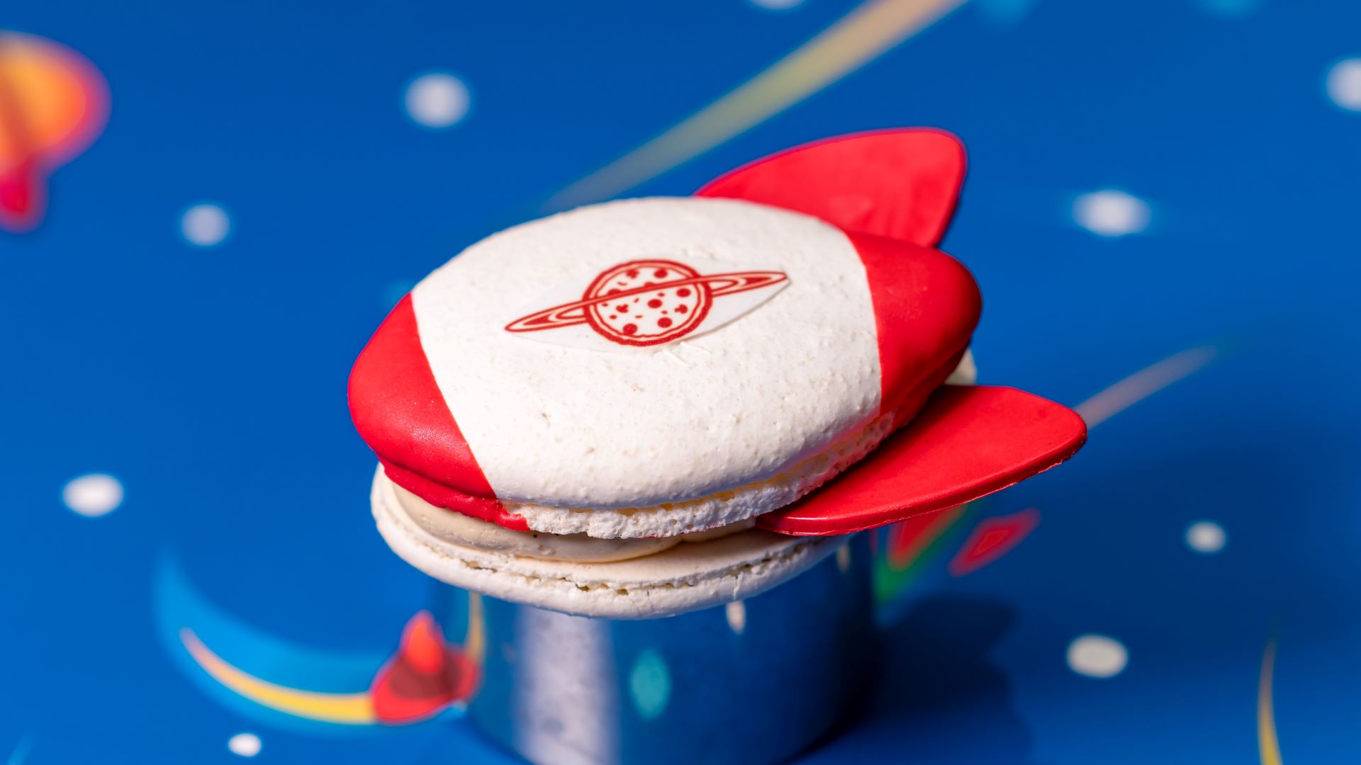 The image features a creatively designed macaron shaped like a rocket, complete with red fins and a logo resembling Saturn on its surface. The macaron is placed on a vibrant blue background decorated with colorful space-themed elements.
