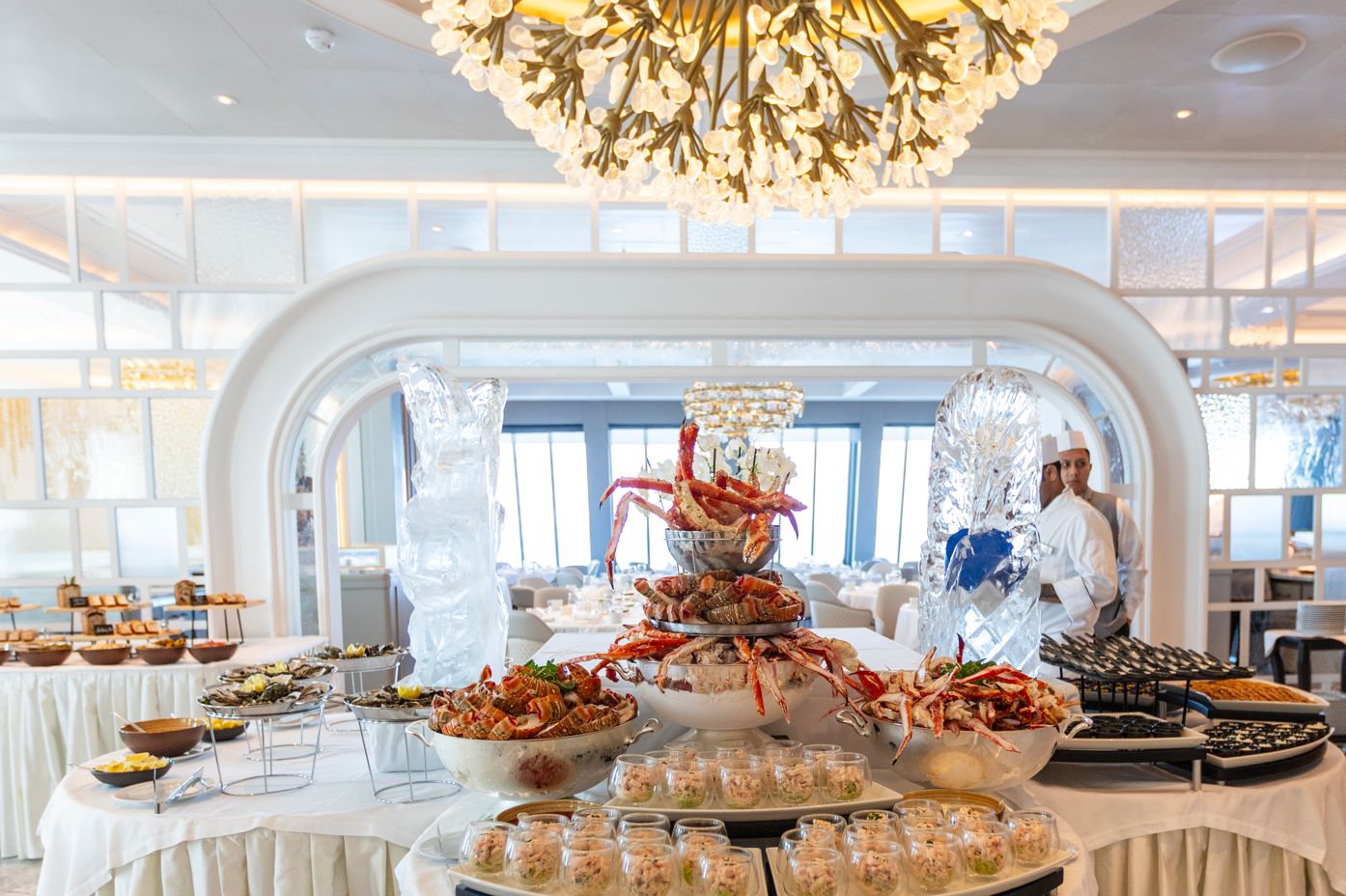The image showcases an elegantly arranged buffet table in a lavish dining setting, featuring an assortment of seafood dishes displayed attractively. In the background, a chef is visible, adding to the sense of culinary sophistication.