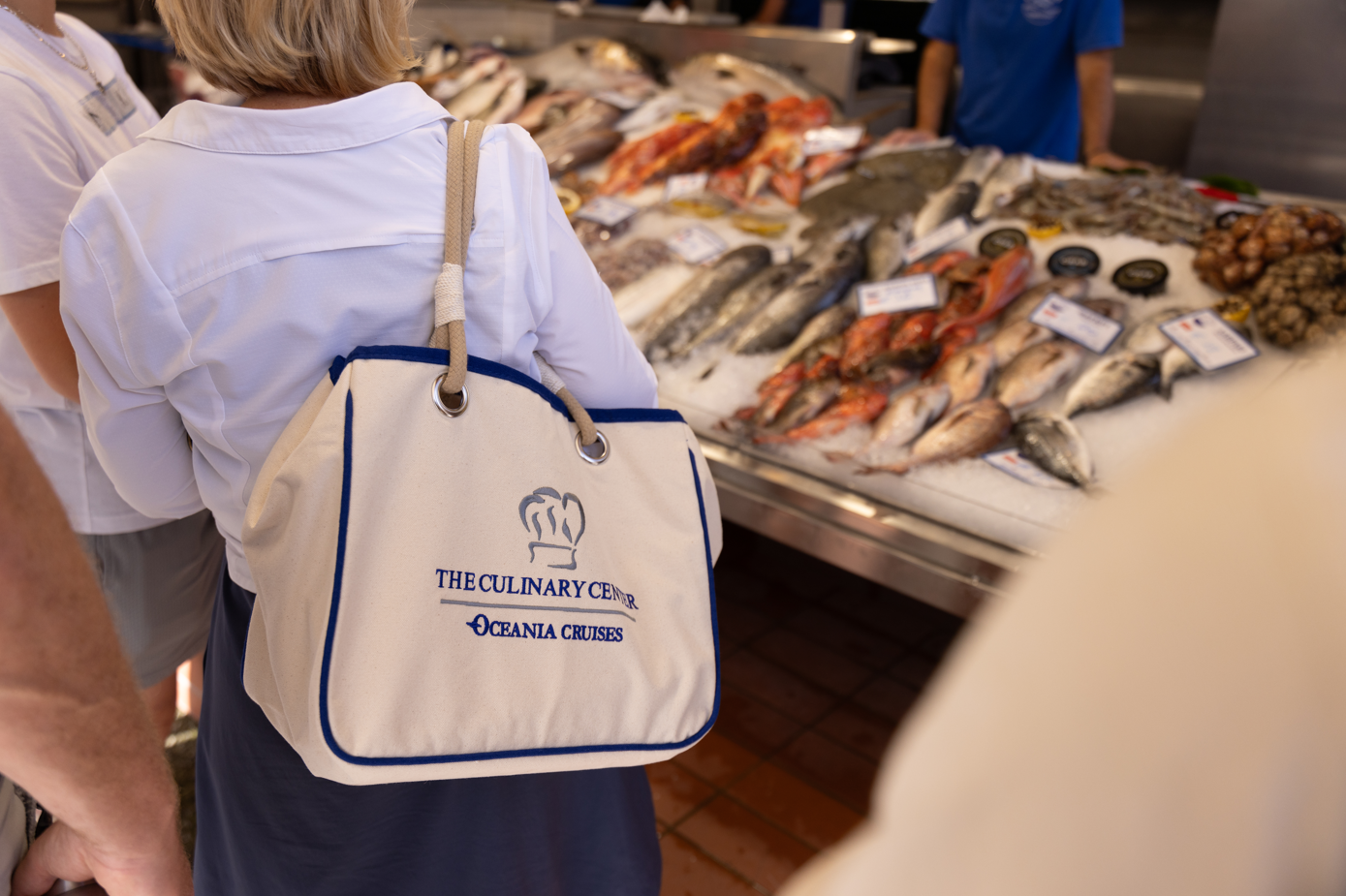 The image shows a woman in a white shirt holding a stylish tote bag while observing a seafood market stall filled with various fish and seafood. The market appears vibrant and well-stocked with a variety of marine products.