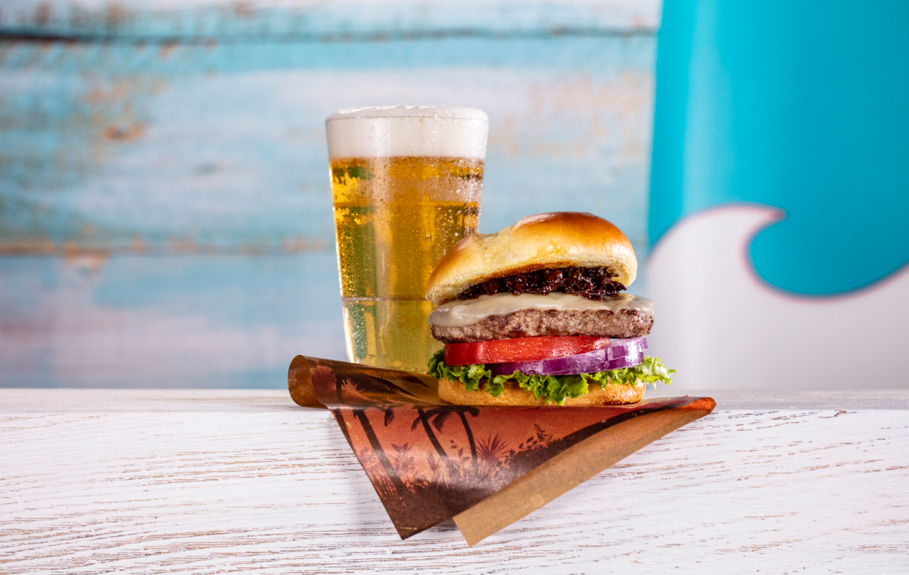 The image features a gourmet hamburger stacked with fresh ingredients, including lettuce, tomato, and onion, alongside a tall glass of golden beer. The setting is casual, with a light blue background that evokes a beach or summer theme.