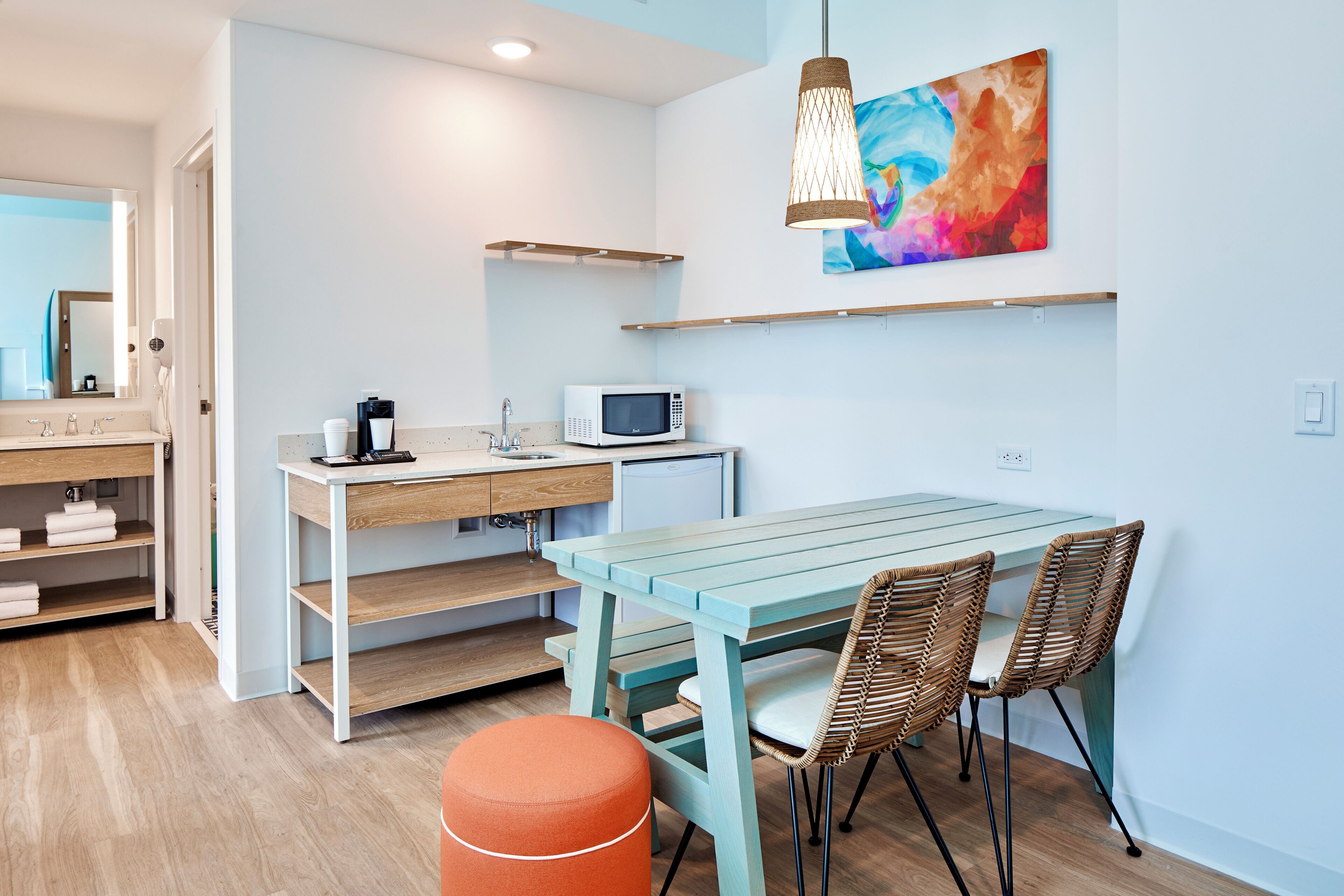 The image depicts a modern kitchenette and dining area within a well-lit interior space. It features a table with chairs, a microwave, and decorative art on the wall.