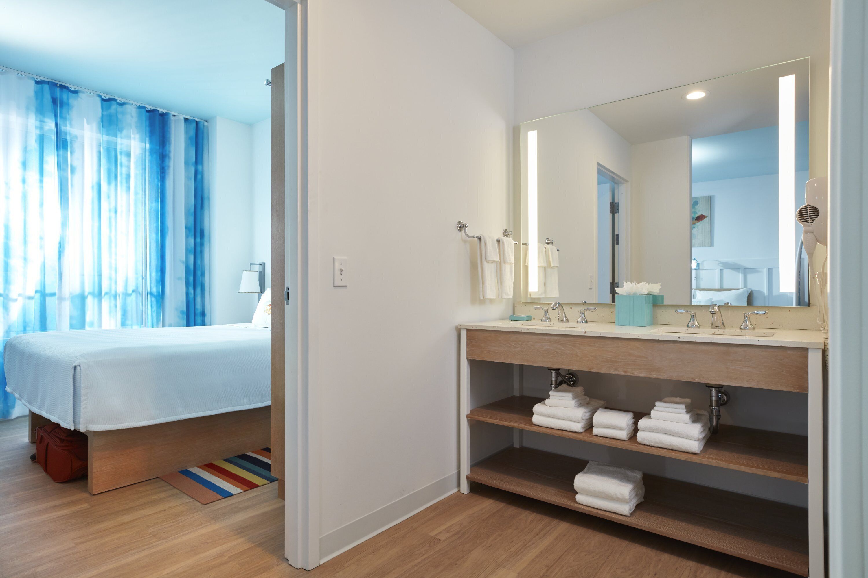 The image shows a well-lit hotel room featuring a neatly made bed and a stylish bathroom area. The bathroom includes a mirror, sink, and neatly stacked towels, contributing to an inviting atmosphere.