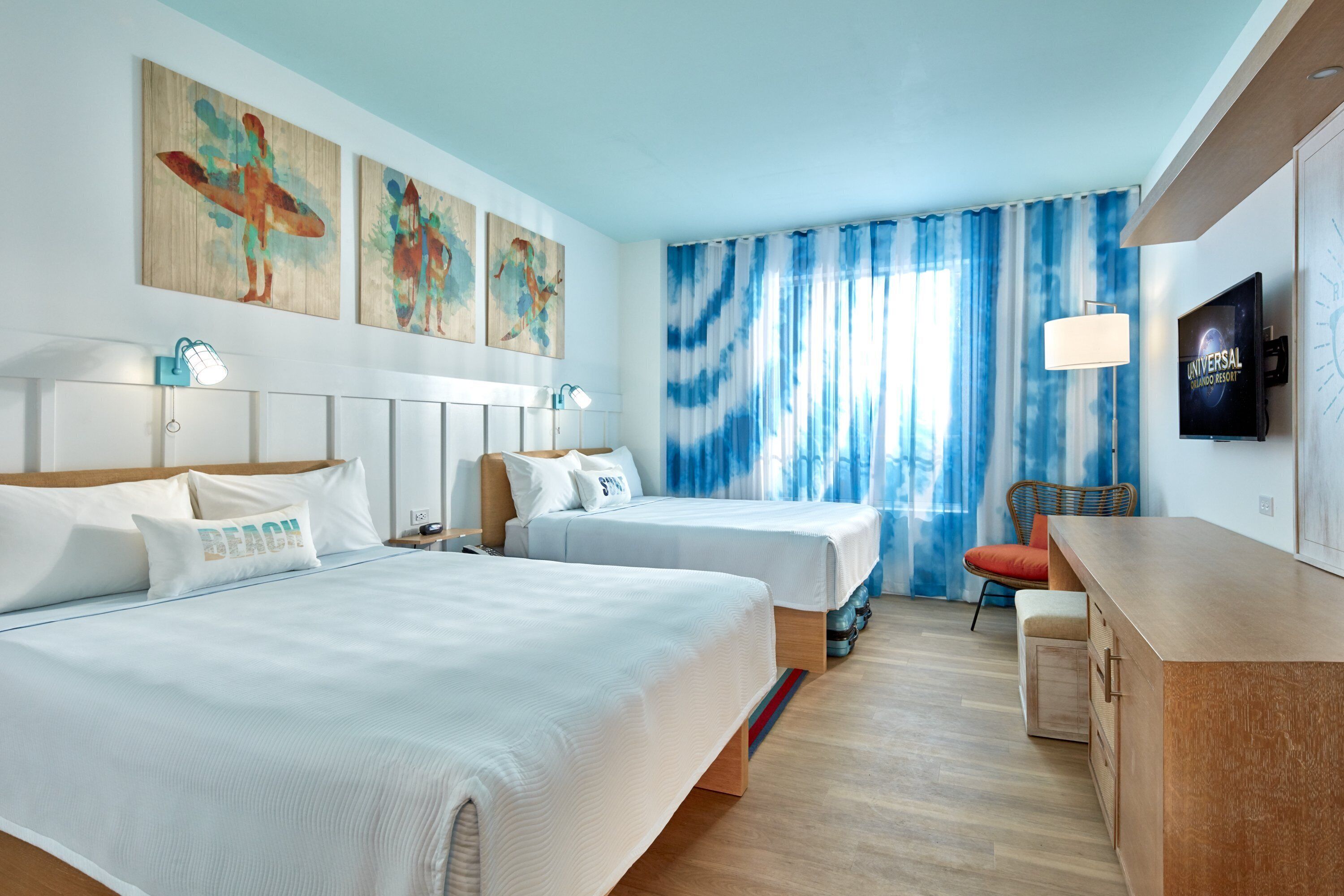 The image shows a brightly lit hotel room featuring two double beds, decorative surfboard artwork, and large windows with blue and white striped curtains. The interior exudes a modern and colorful beach theme.