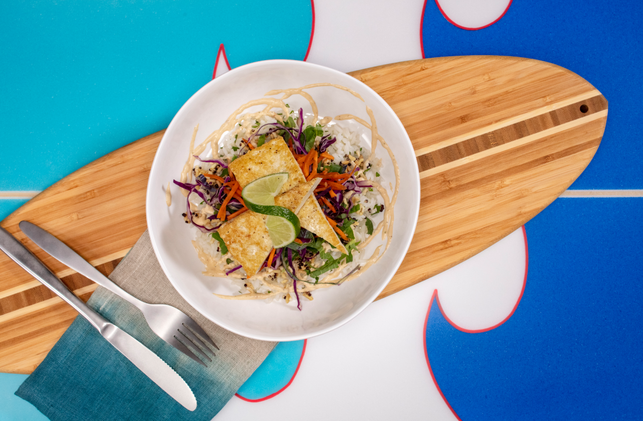 A visually appealing bowl of vibrant salad topped with a golden piece of grilled fish is presented on a wooden surfboard. The colorful ingredients include shredded purple cabbage, carrots, and fresh herbs, creating an inviting dish with a tropical aesthetic.