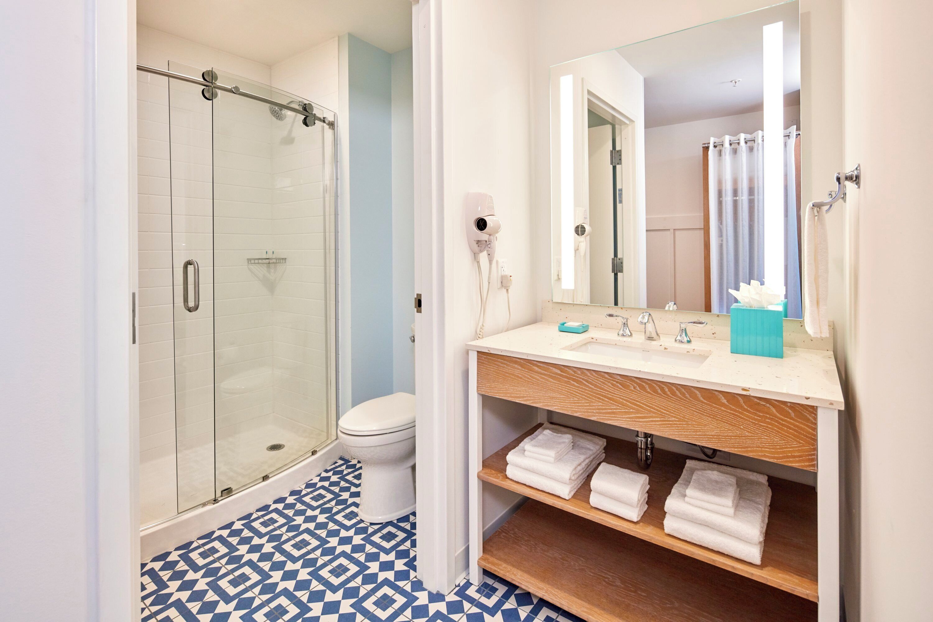 A modern bathroom featuring a shower area with clear glass doors, a wooden vanity with neatly stacked towels, and a mirror above the sink. The floor is decorated with intricate blue and white patterned tiles.
