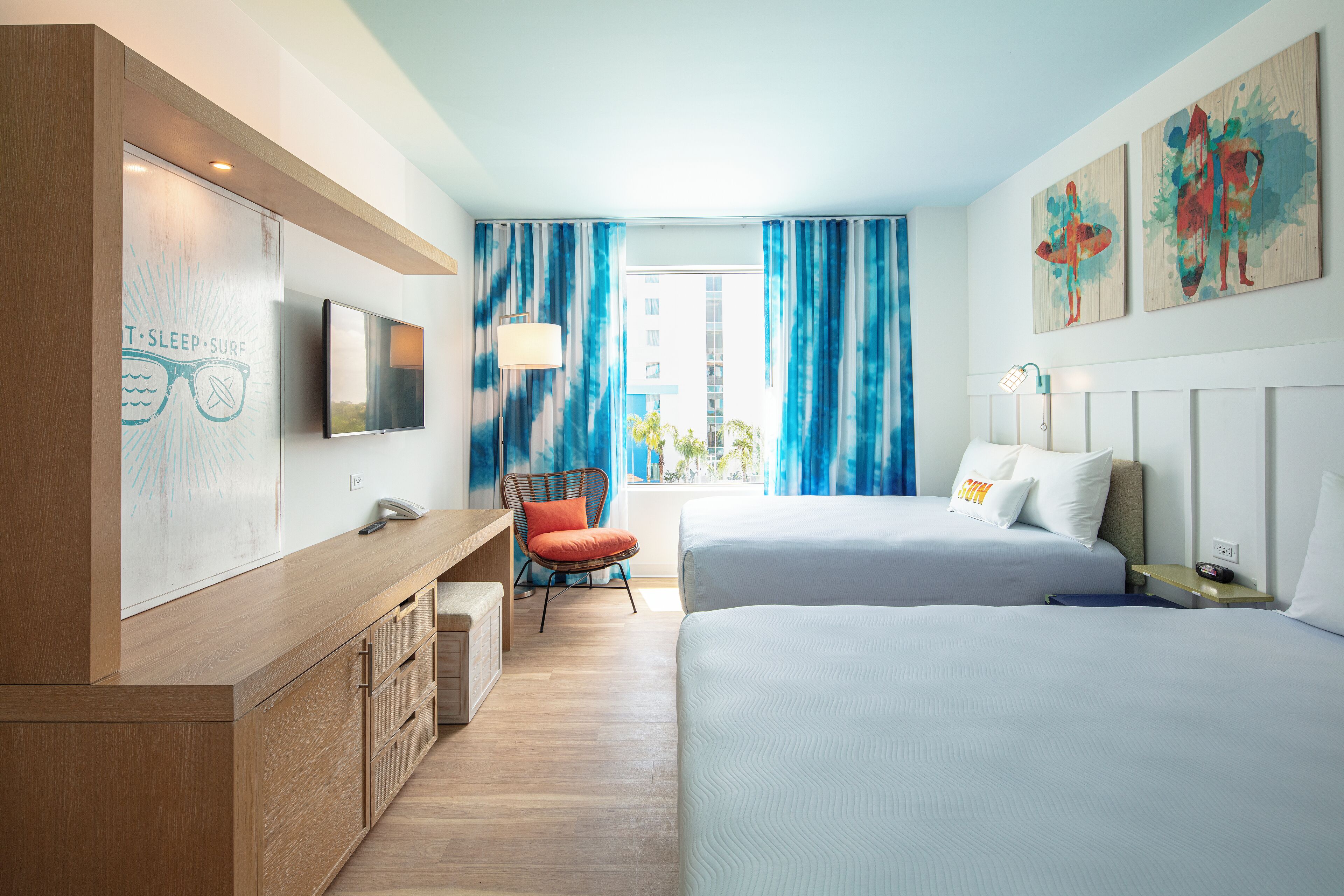 A brightly lit hotel room featuring two beds with blue bedding, a wooden accent wall, and a stylish seating area with a red chair. The room has large windows adorned with blue and white curtains, providing a view of the outside.