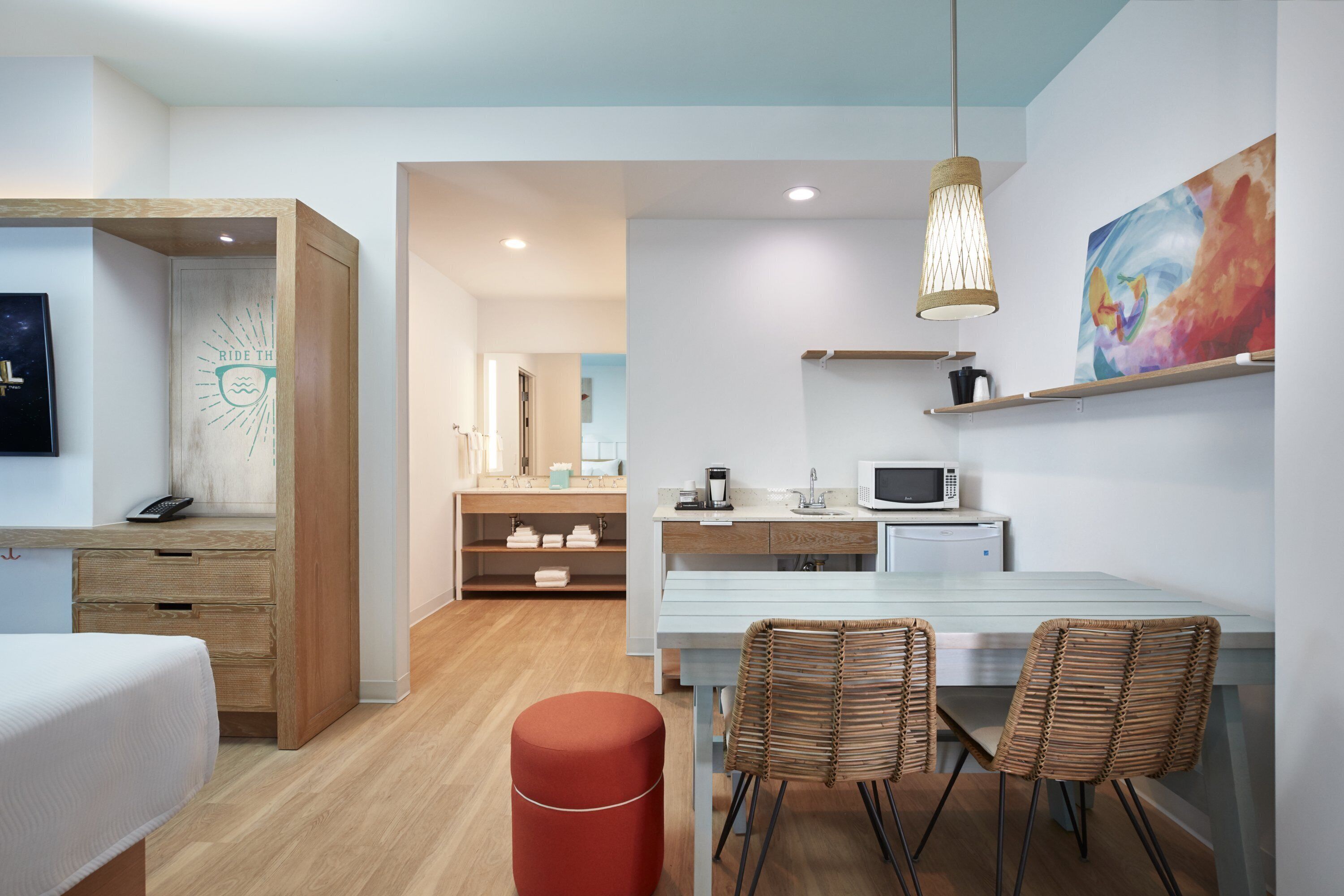 The image depicts a modern hotel room featuring a combined living and dining space. It includes a kitchenette with a coffee maker and a dining table with woven chairs.