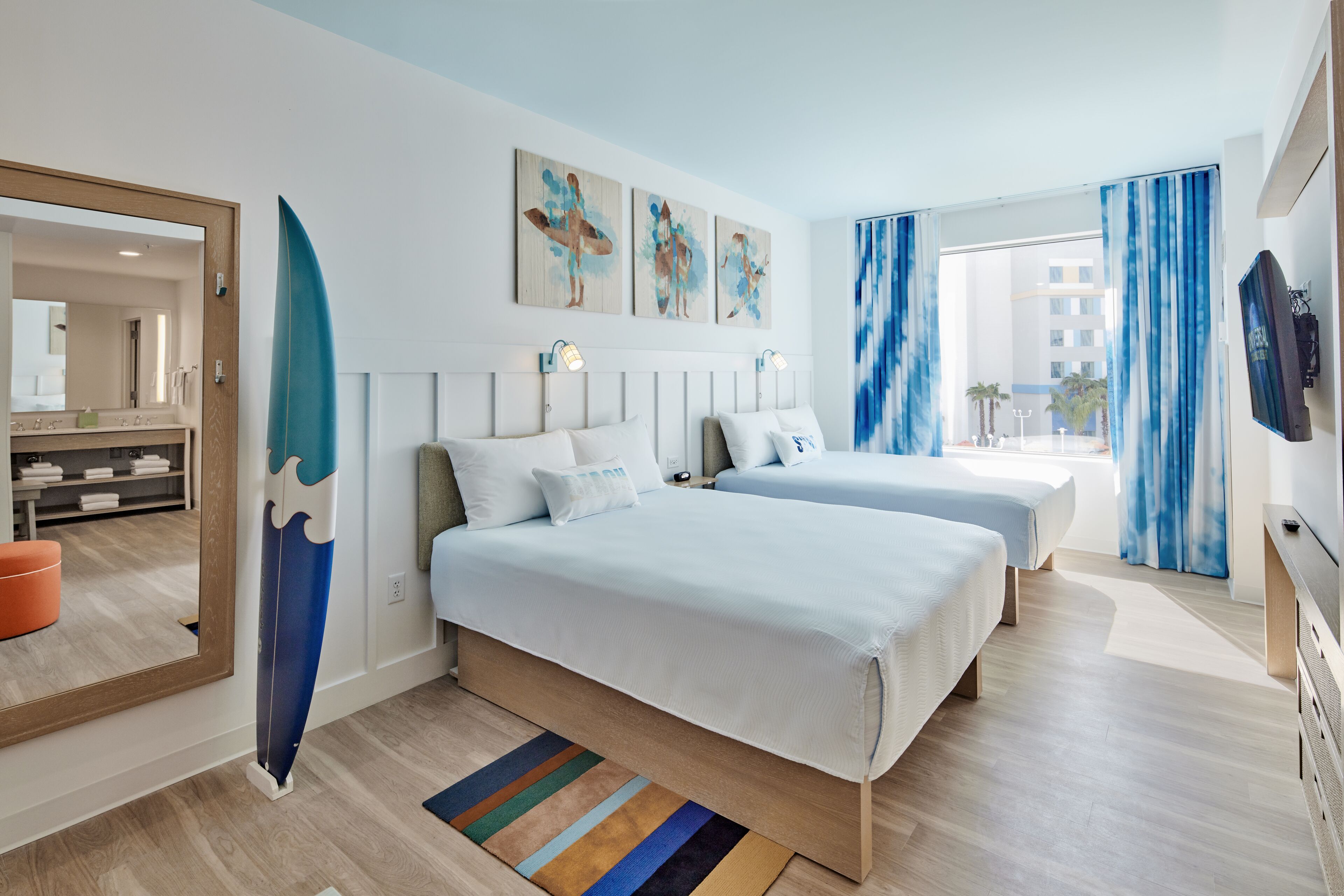 The image showcases a bright and modern hotel room featuring two beds, a large window with sheer curtains, and ocean-themed artwork on the walls. A surfboard decorates the room, adding to the coastal vibe.