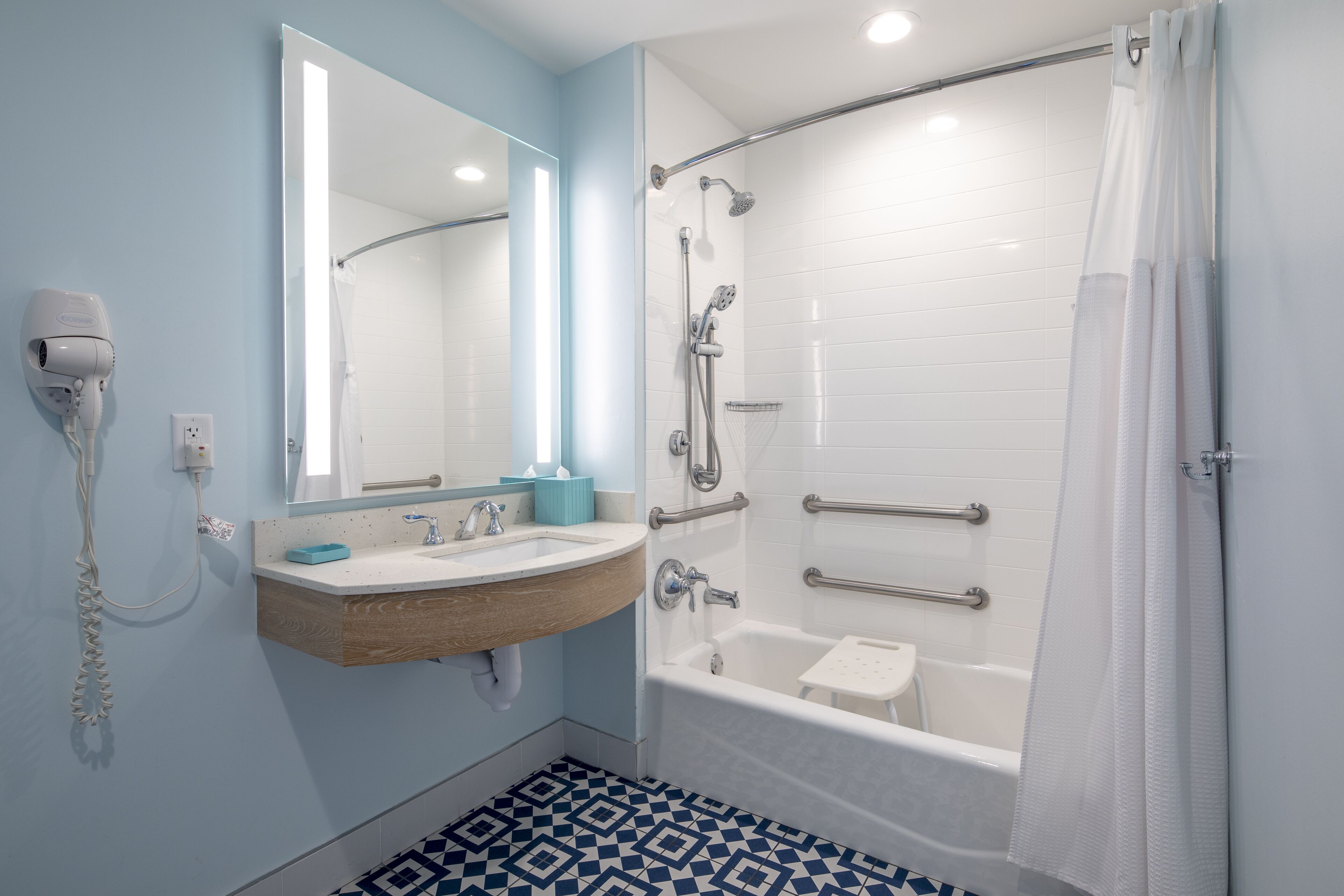The image shows a modern bathroom with a shower and bathtub combination. It features a light blue wall, a large mirror, and safety grab bars, indicating an accessibility-friendly design.
