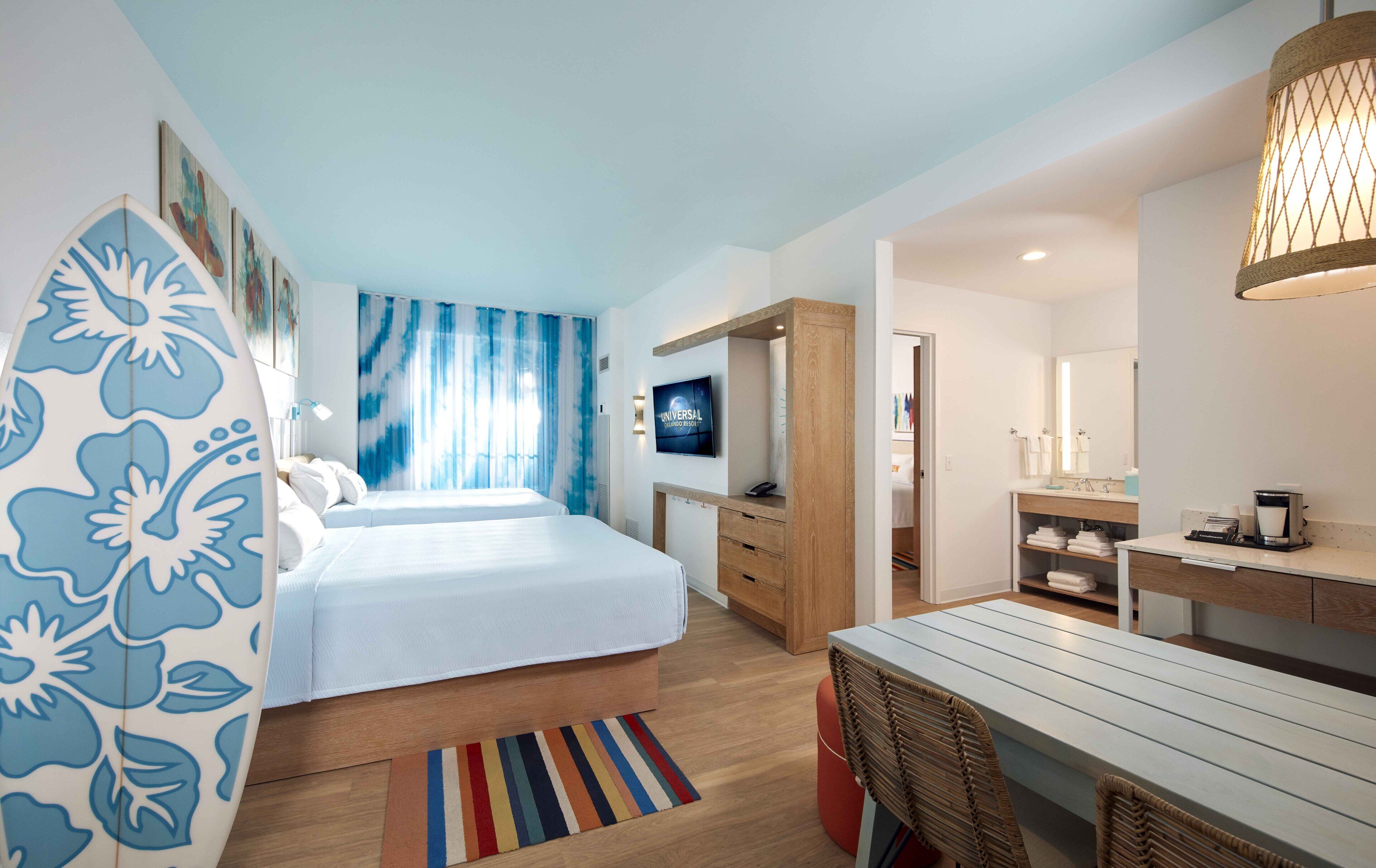 The image features a modern hotel room with two beds, a vibrant blue decor, and a cozy seating area. The design includes tropical-themed artwork and a surfboard decoration, suggesting a beach or resort setting.
