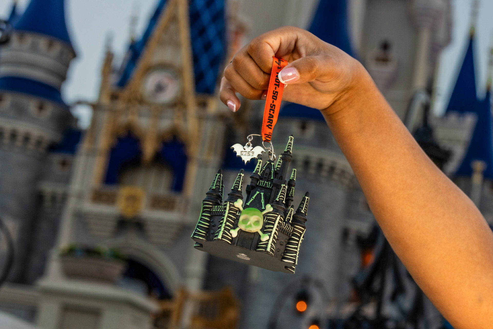 A person holds up a Halloween-themed castle ornament in front of an iconic castle backdrop. The ornament features spooky designs, including a skull, and is part of a seasonal collection.
