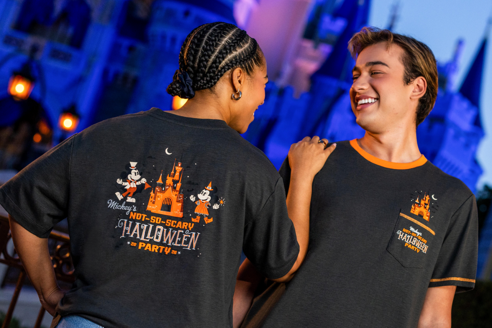 The image features two individuals at a themed event, wearing matching Halloween party T-shirts. The backdrop includes a castle, suggesting a festive atmosphere at a theme park.
