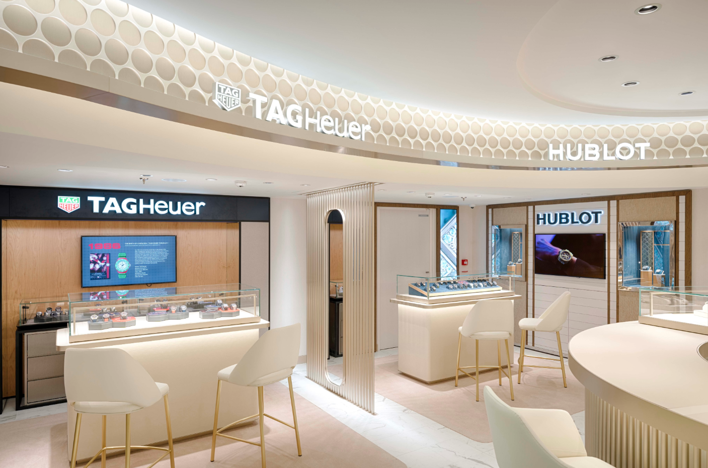 The image depicts a luxury watch retail store interior featuring displays for TAG Heuer and Hublot. The design is modern with elegant fixtures and a neutral color palette, showcasing various watch models in glass cases.