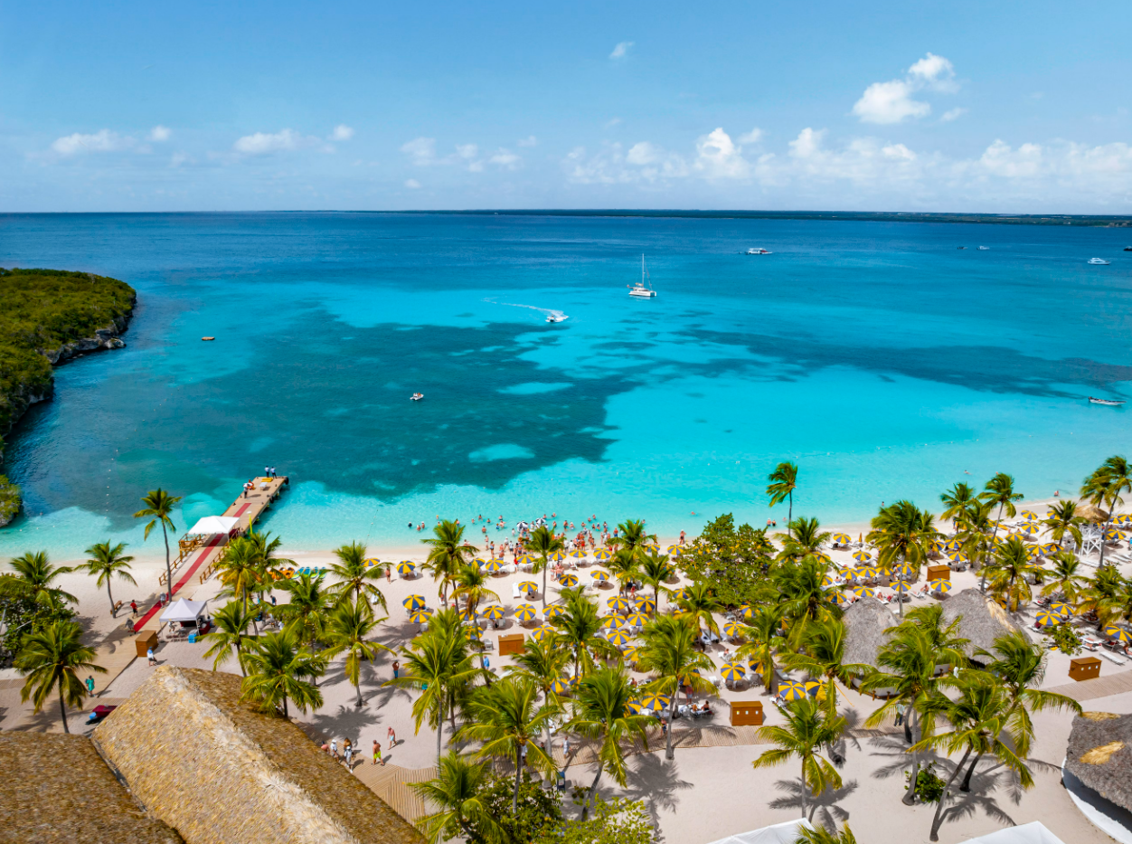 A panoramic view of a tropical beach showcasing clear turquoise waters and a sandy shoreline dotted with beach umbrellas and lounge chairs. The scene is framed by lush green palm trees and features a dock extending into the water.