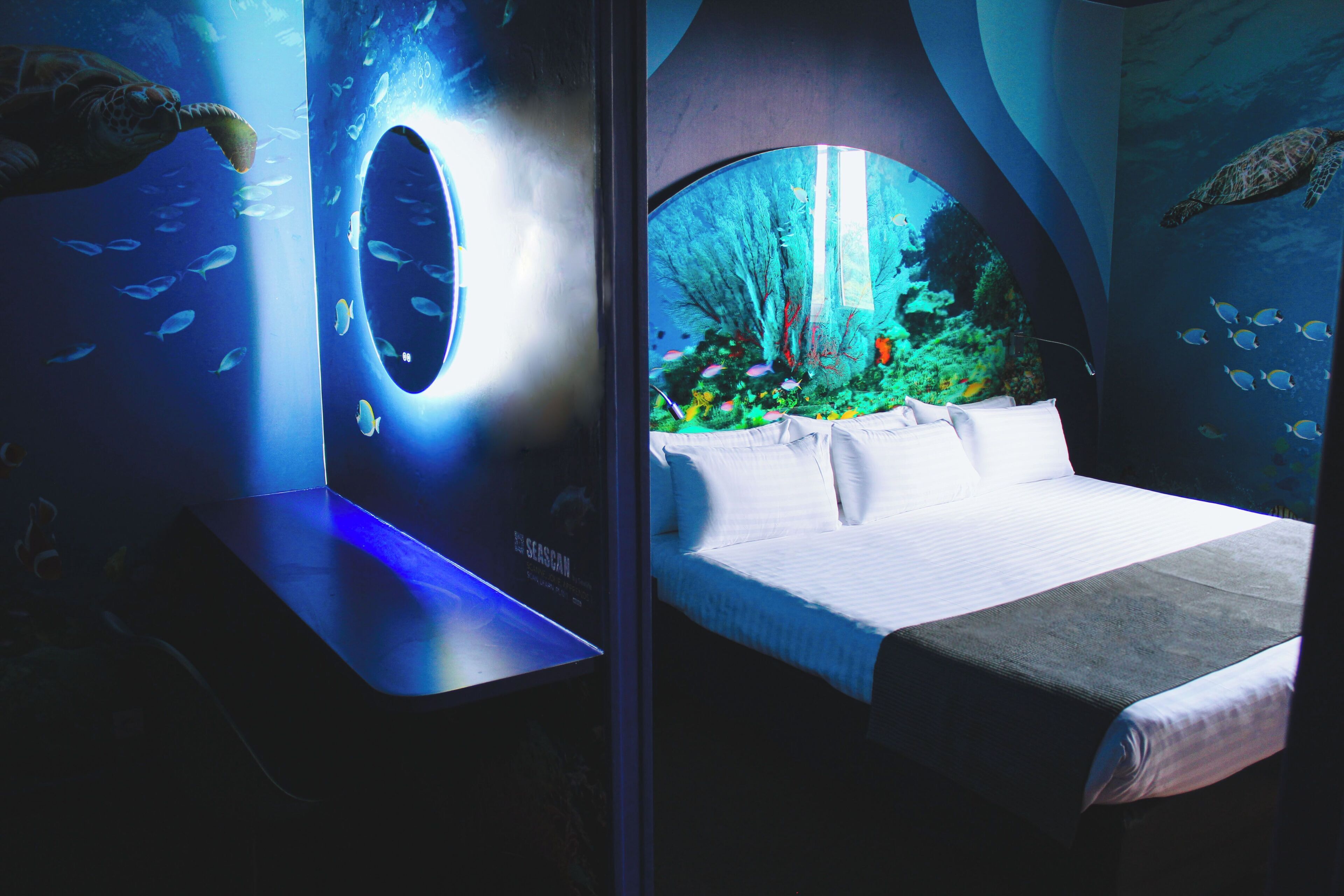 The image showcases an imaginative hotel room decorated with an underwater theme. The room features a bed with white linens and a vibrant mural of marine life on the wall, complemented by a circular mirror that emits a soft blue light.