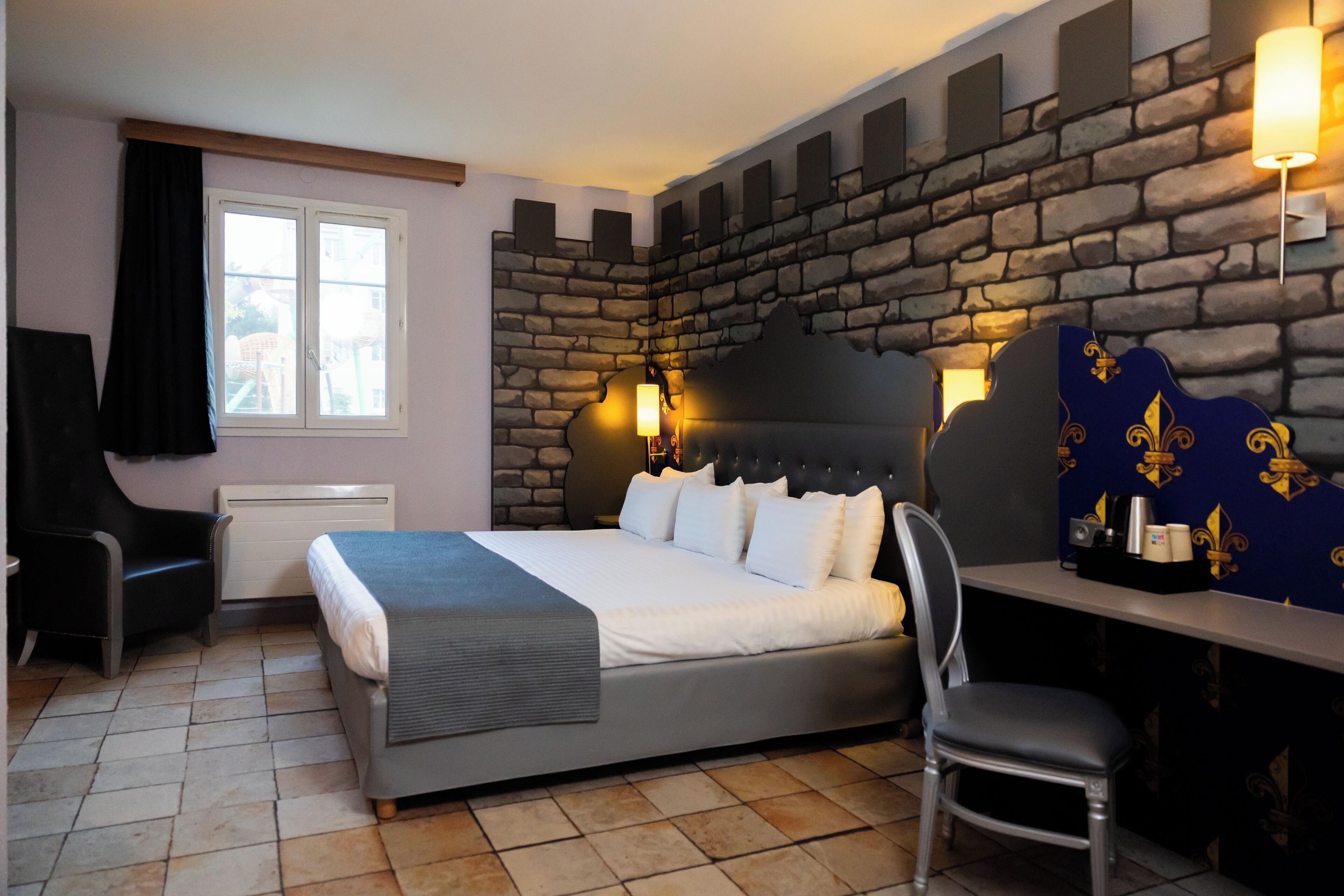 The image depicts a well-furnished hotel room featuring a modern design with a unique castle-like decor on the walls. The room includes a large bed with white linens, a decorative chair, and a desk area.