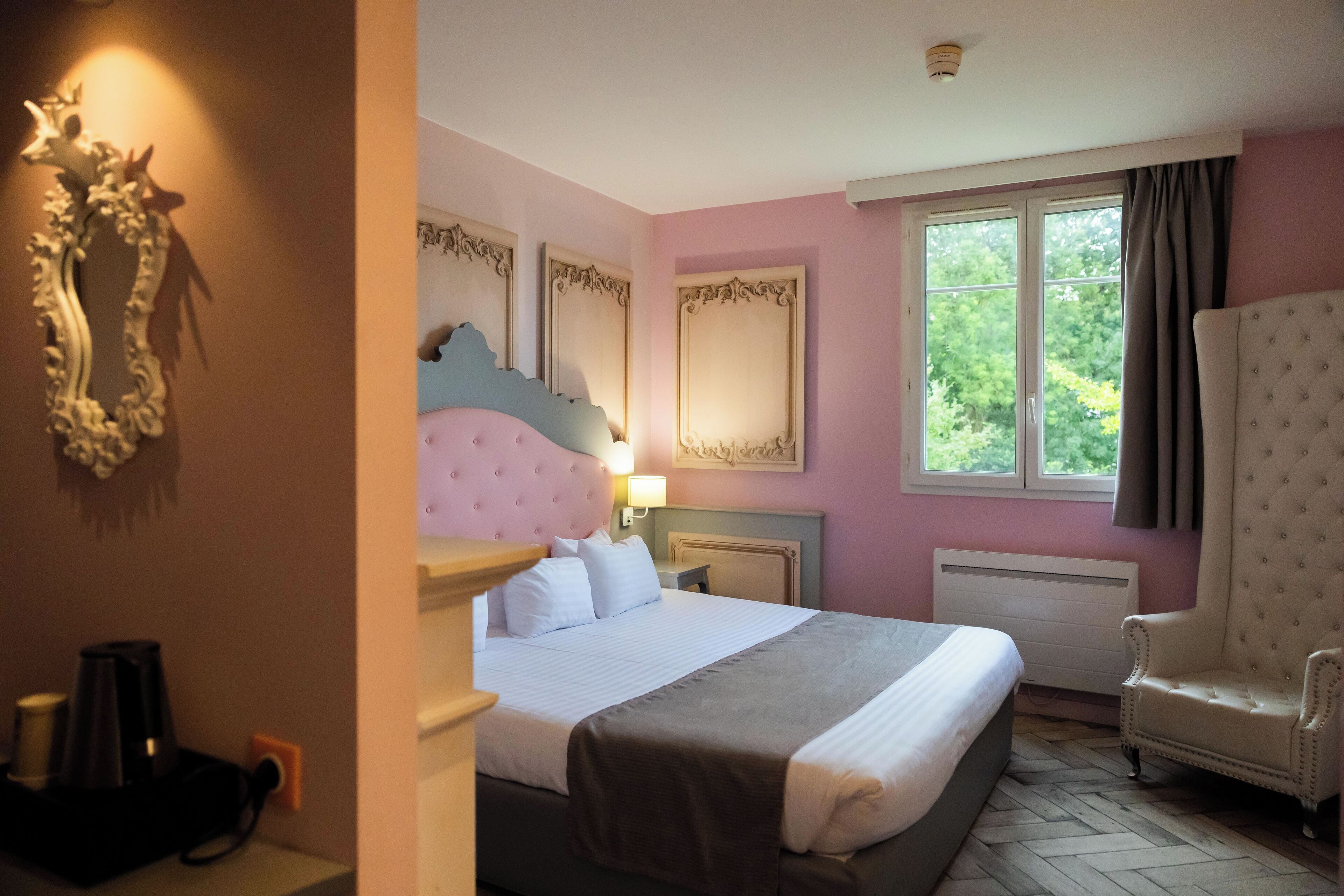 A cozy hotel room featuring a queen-sized bed with a light gray blanket and decorative pillows. The walls are painted in soft pink, complemented by a large, ornate white mirror and a stylish wingback chair, with a window providing natural light and a view of greenery outside.
