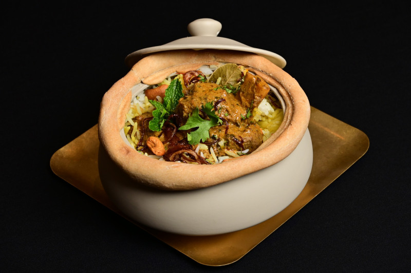 The image features a traditional dish served in a decorative pot, showcasing a flavorful biryani with meat, garnished with herbs and spices. The presentation emphasizes the culinary artistry of the dish, set against a dark background.