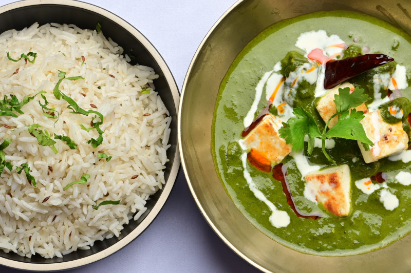 The image features a traditional Indian meal consisting of a bowl of palak paneer, a spinach-based curry with paneer cheese cubes, accompanied by a serving of steamed rice. The dish is garnished with a swirl of cream, green herbs, and red chili, presented in a decorative metal bowl.