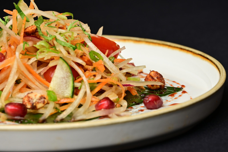 The image features a vibrant, colorful salad dish artistically arranged on a white plate. The salad includes a mix of shredded vegetables, nuts, and garnishes, set against a dark background, highlighting its freshness and appeal.