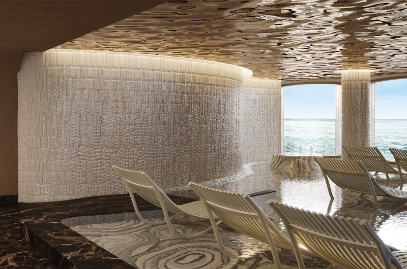 The image showcases a modern indoor relaxation area featuring lounge chairs arranged beside a tranquil water feature. Large windows provide a view of the ocean, creating a serene atmosphere enhanced by soft lighting and contemporary design elements.
