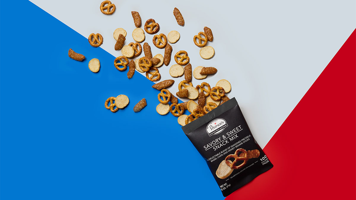 The image features a bag of snack mix spilling onto a colored background split into blue, white, and red sections. The mix includes various types of pretzels and crackers.