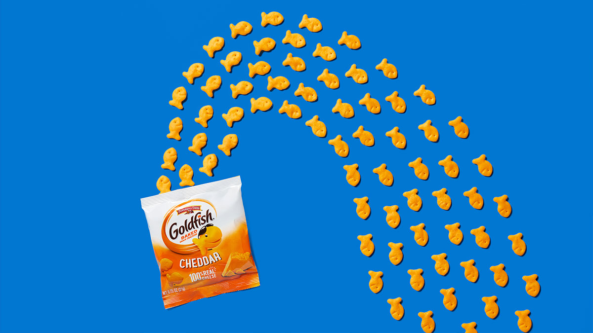 The image features a vibrant blue background with numerous small goldfish-shaped crackers arranged in an arch. At the bottom left, there's a package of Goldfish Cheddar crackers prominently displayed.
