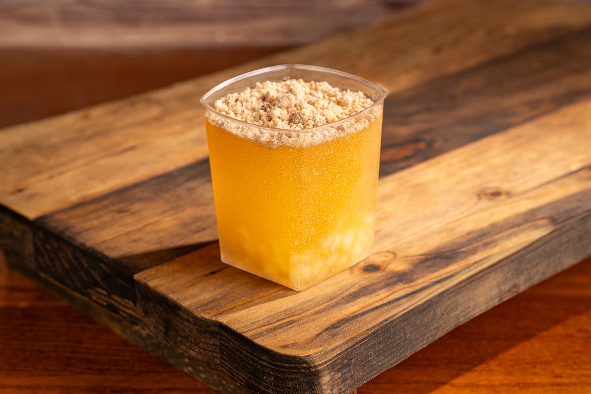 The image showcases a clear plastic cup filled with a yellow-colored beverage topped with a layer of crushed ingredients. The setting features a wooden surface, adding a rustic touch to the presentation.