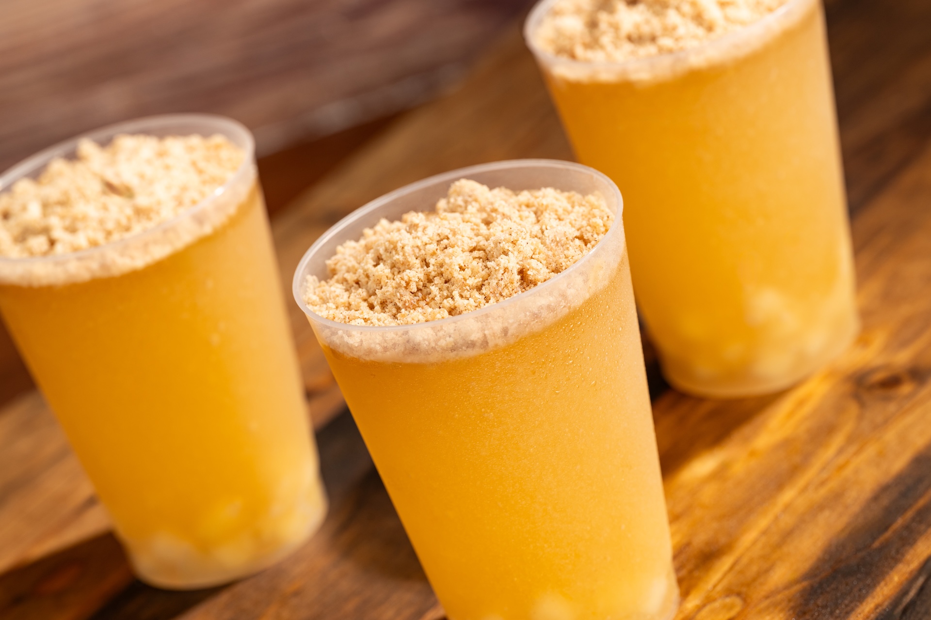 The image features three transparent cups filled with a frosty yellow beverage topped with a crumbly beige mixture. The setting suggests a casual food or drink event, highlighting refreshing summer treats.