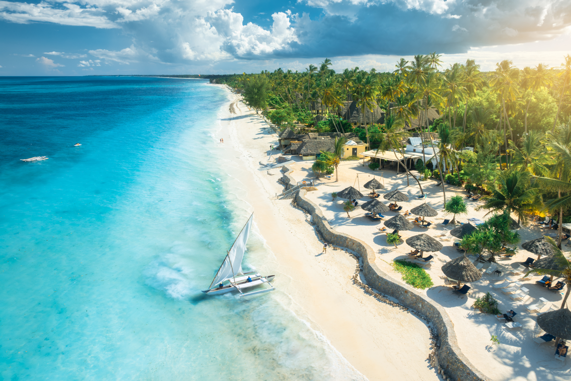 A picturesque aerial view of a sandy beach along a tropical coastline. The scene features clear turquoise waters, a sailboat gliding through the waves, and a lush landscape dotted with palm trees and thatched-roof structures.