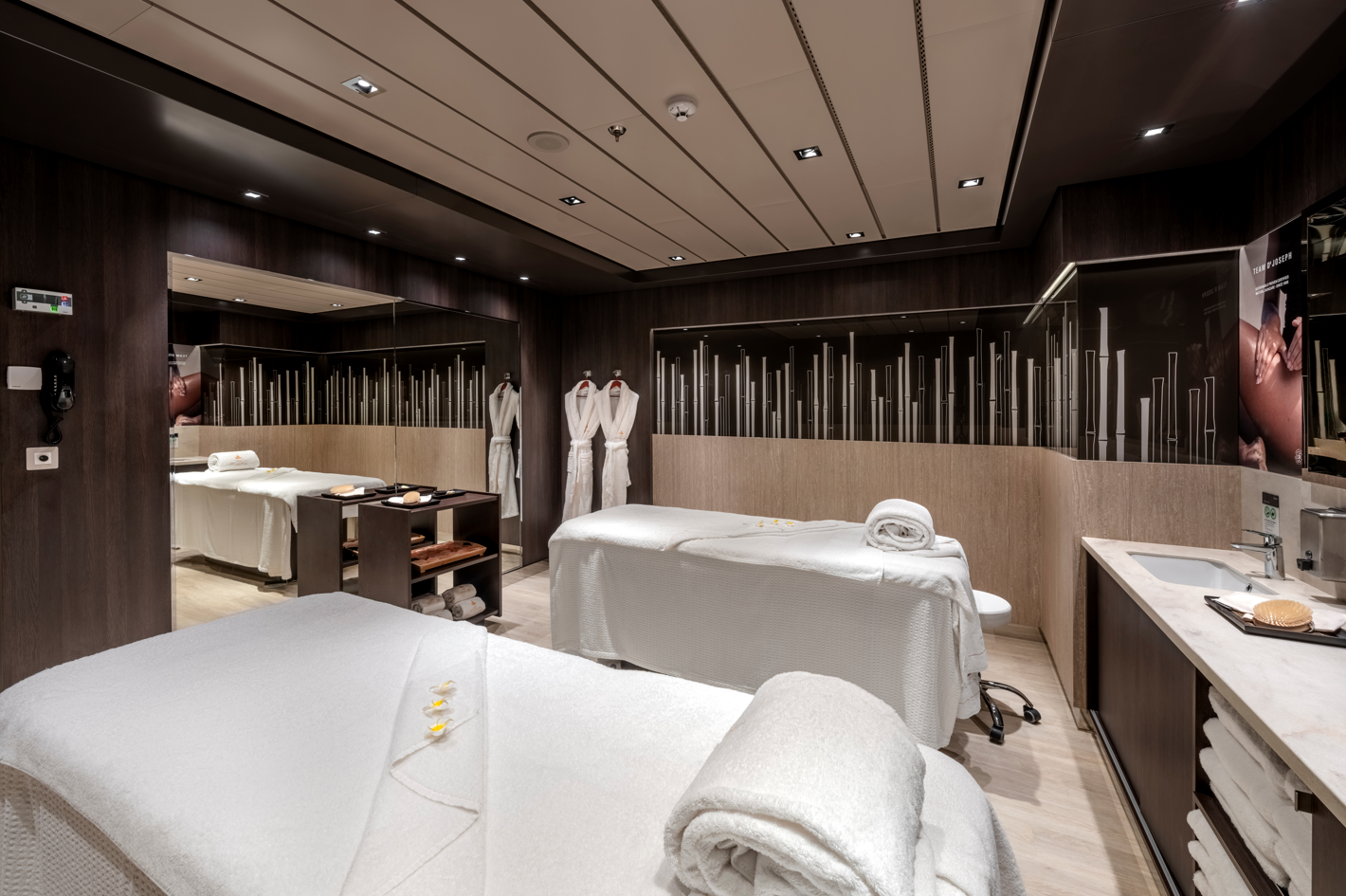 The image depicts a luxurious spa treatment room featuring two massage beds, elegantly arranged with soft white linens. The modern interior design includes wood paneling and subtle decorative accents, creating a serene and calming atmosphere.