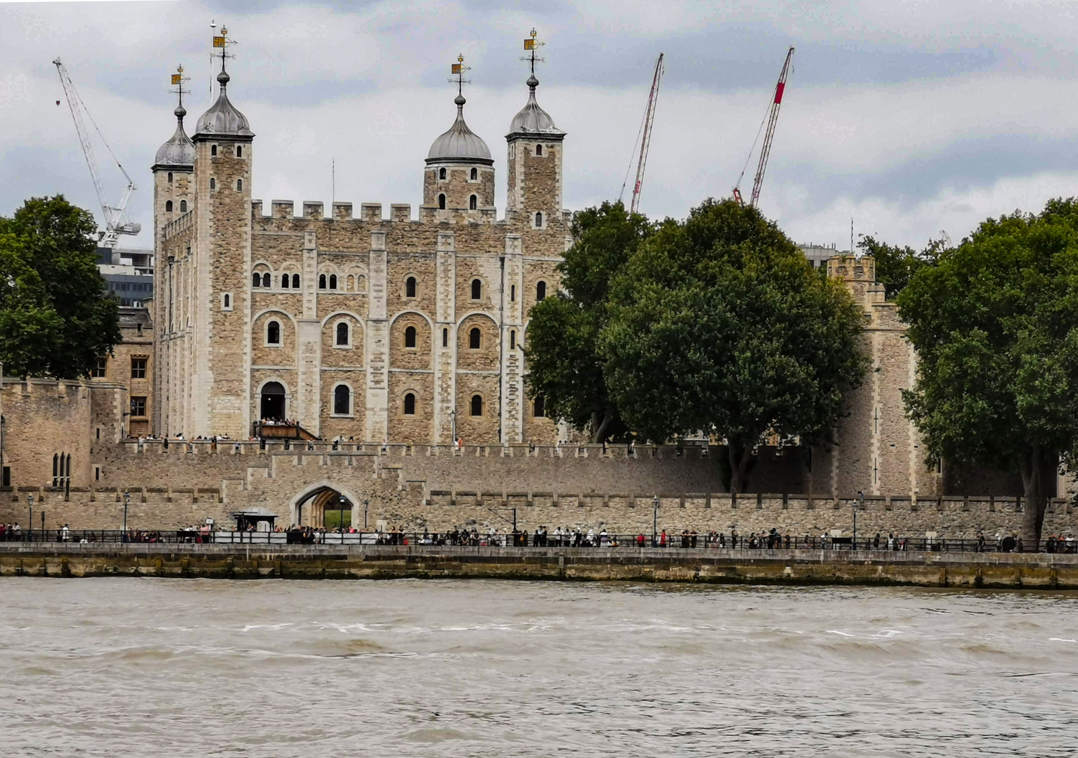 The image shows the Tower of London, a historic castle located on the north bank of the River Thames in central London. The castle features stone walls, several towers, and is surrounded by lush greenery, with people visible along the riverbank.