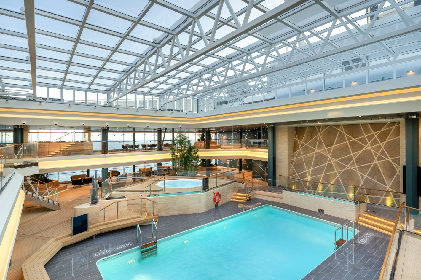 The image showcases a modern indoor swimming pool area featuring a glass ceiling that allows natural light to illuminate the space. The design includes wooden accents, stylish seating areas, and a geometric stone wall, creating an inviting atmosphere.