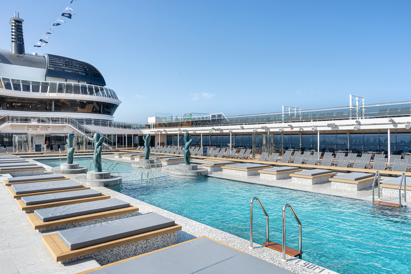 The image depicts a luxurious cruise ship pool area featuring a pristine swimming pool surrounded by sun loungers. Elegant sculptures of figures adorn the pool, and the ship's upper deck showcases a clear blue sky and sea in the background.