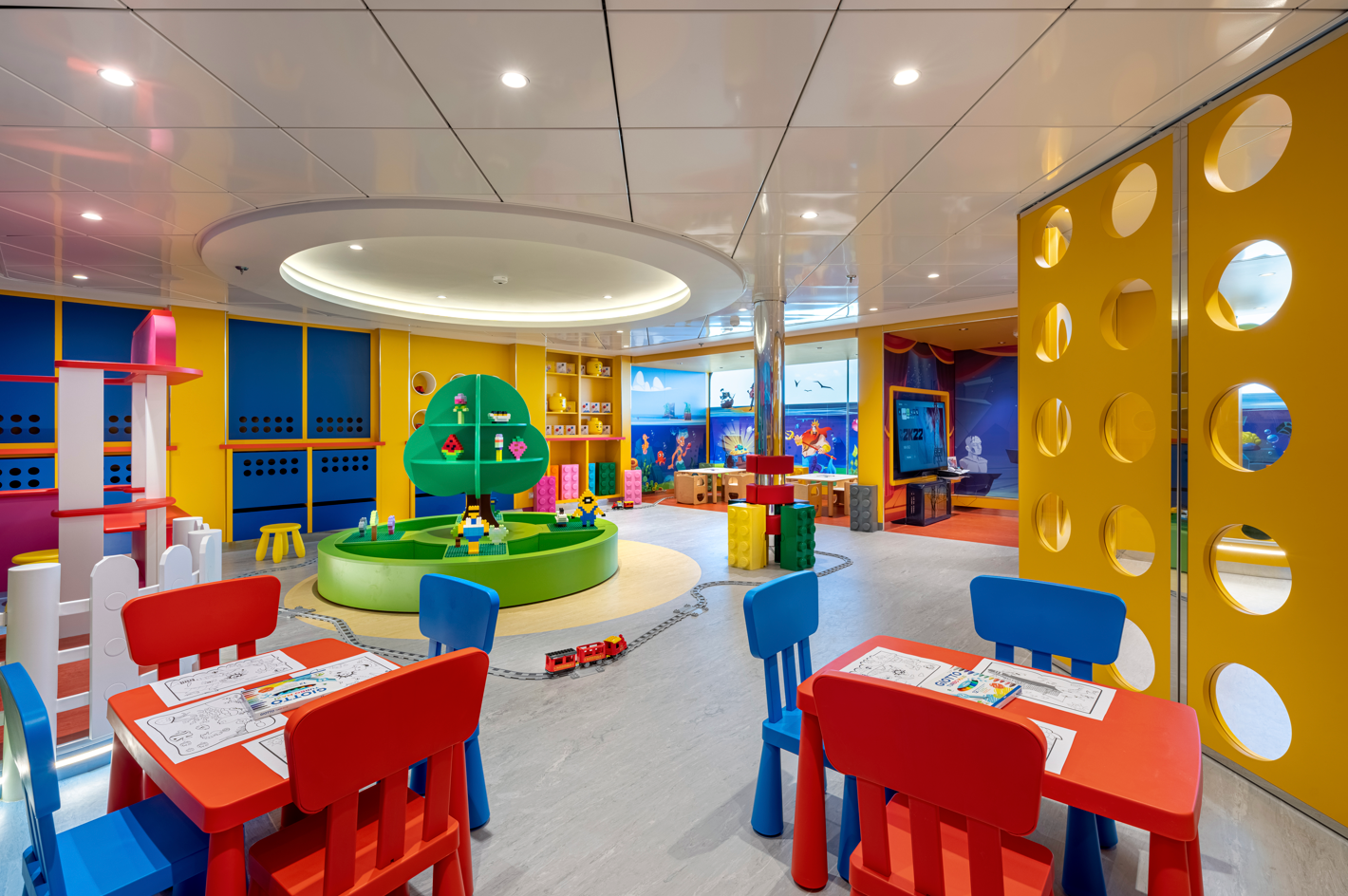 The image depicts a brightly colored children's playroom featuring a circular play area with interactive toys and games. The room includes various seating arrangements with red, blue, and yellow furniture, creating a vibrant and welcoming environment for young children.