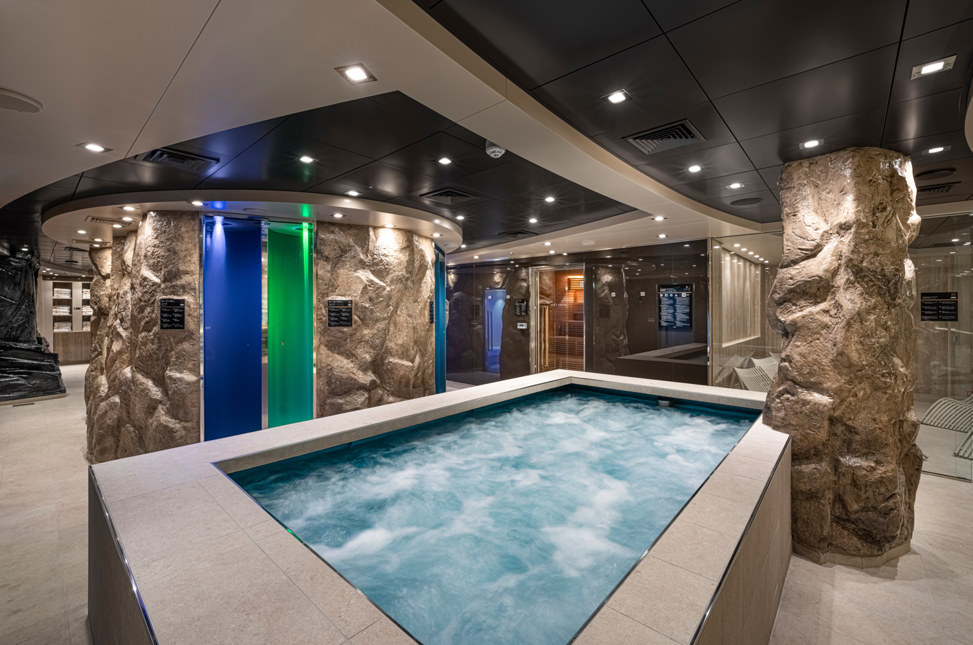 The image showcases a luxurious spa area featuring a stone hot tub filled with bubbling water. The modern interior includes ambient lighting, decorative stone walls, and various spa facilities in a serene setting.