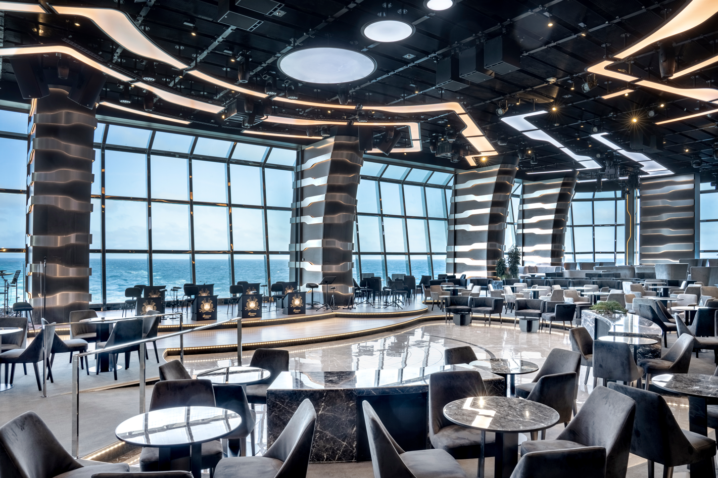 The image depicts the interior of a modern restaurant or lounge with a panoramic view of the ocean. The space features stylish decor, including wavy lighting designs, elegant seating arrangements, and large windows that allow natural light to fill the space.
