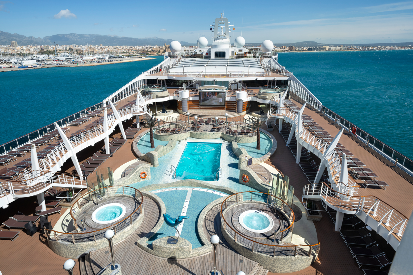 The image showcases the top deck of a cruise ship featuring several pools and hot tubs. The deck is surrounded by sun loungers and offers a panoramic view of the ocean and coastline.