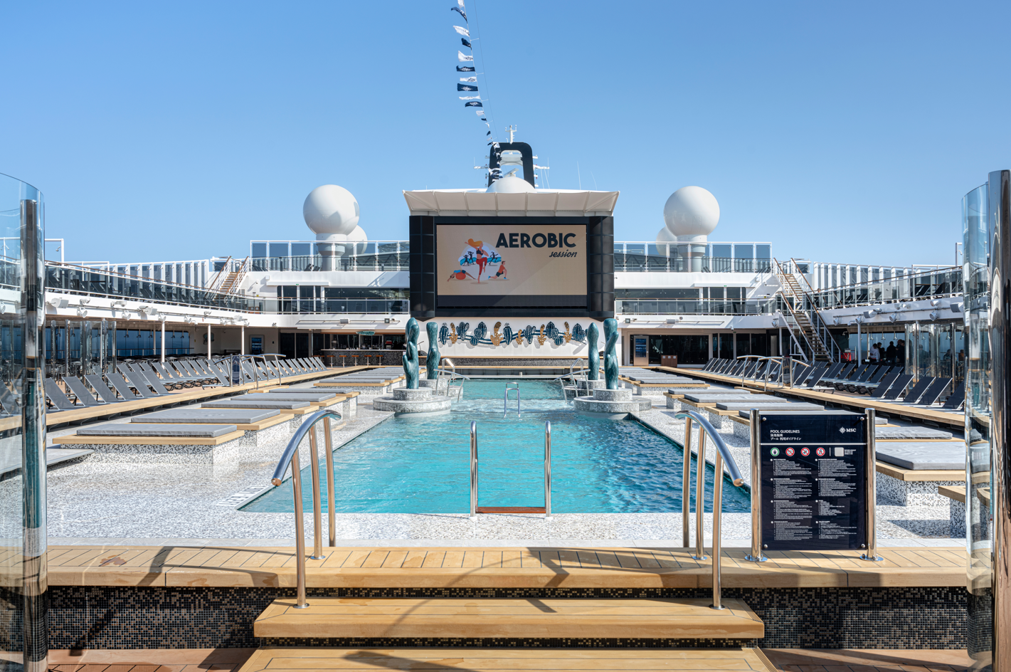 The image depicts a modern cruise ship's pool deck, showcasing a large swimming pool surrounded by lounge chairs and a giant screen displaying promotional content for 'Aerobic.' The setting is bright and sunny, typical of a leisure environment.
