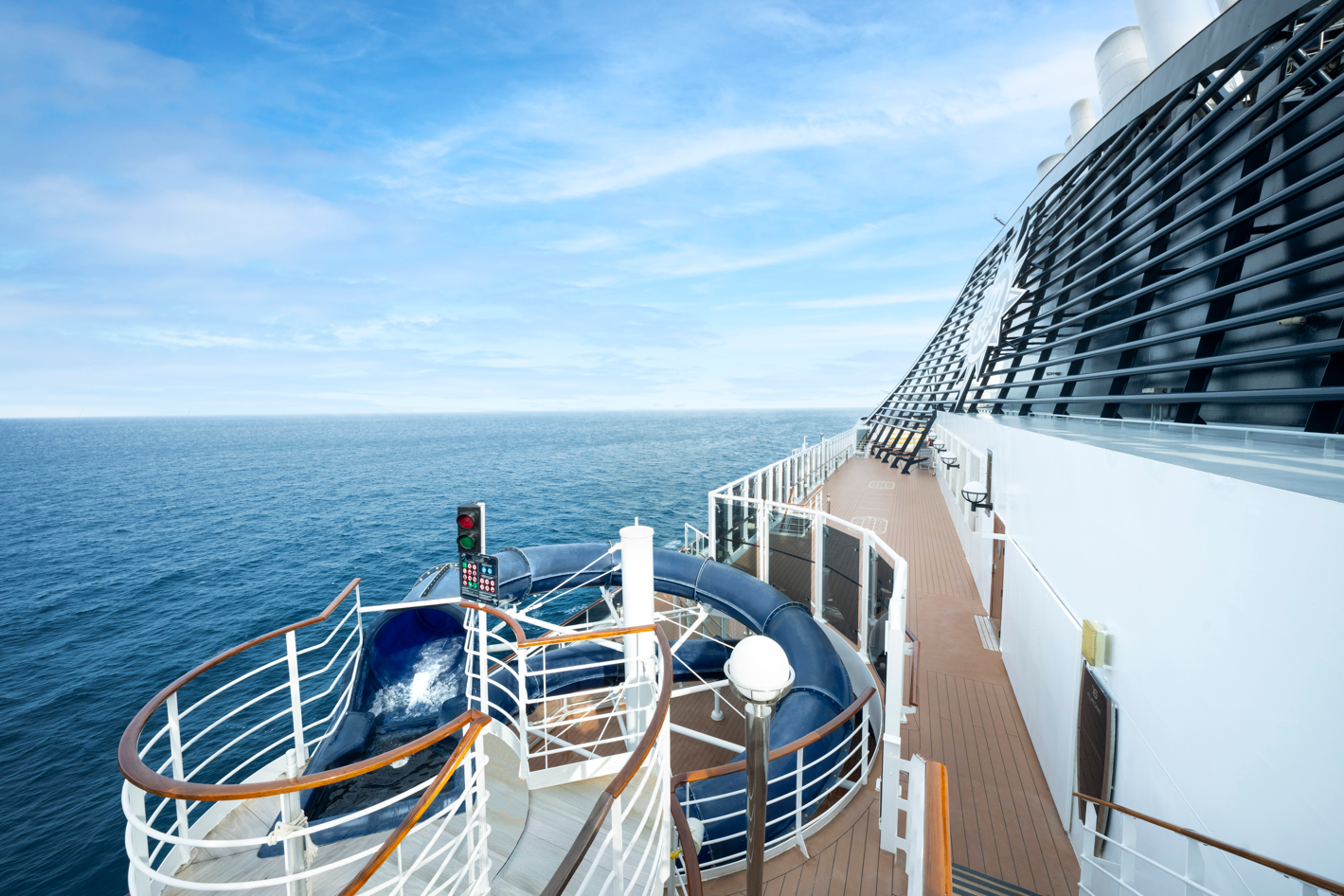 A view from the deck of a cruise ship overlooking the open ocean. The image captures a waterslide and safety signals positioned beside the ship's railing, with calm waters stretching into the horizon under a clear sky.